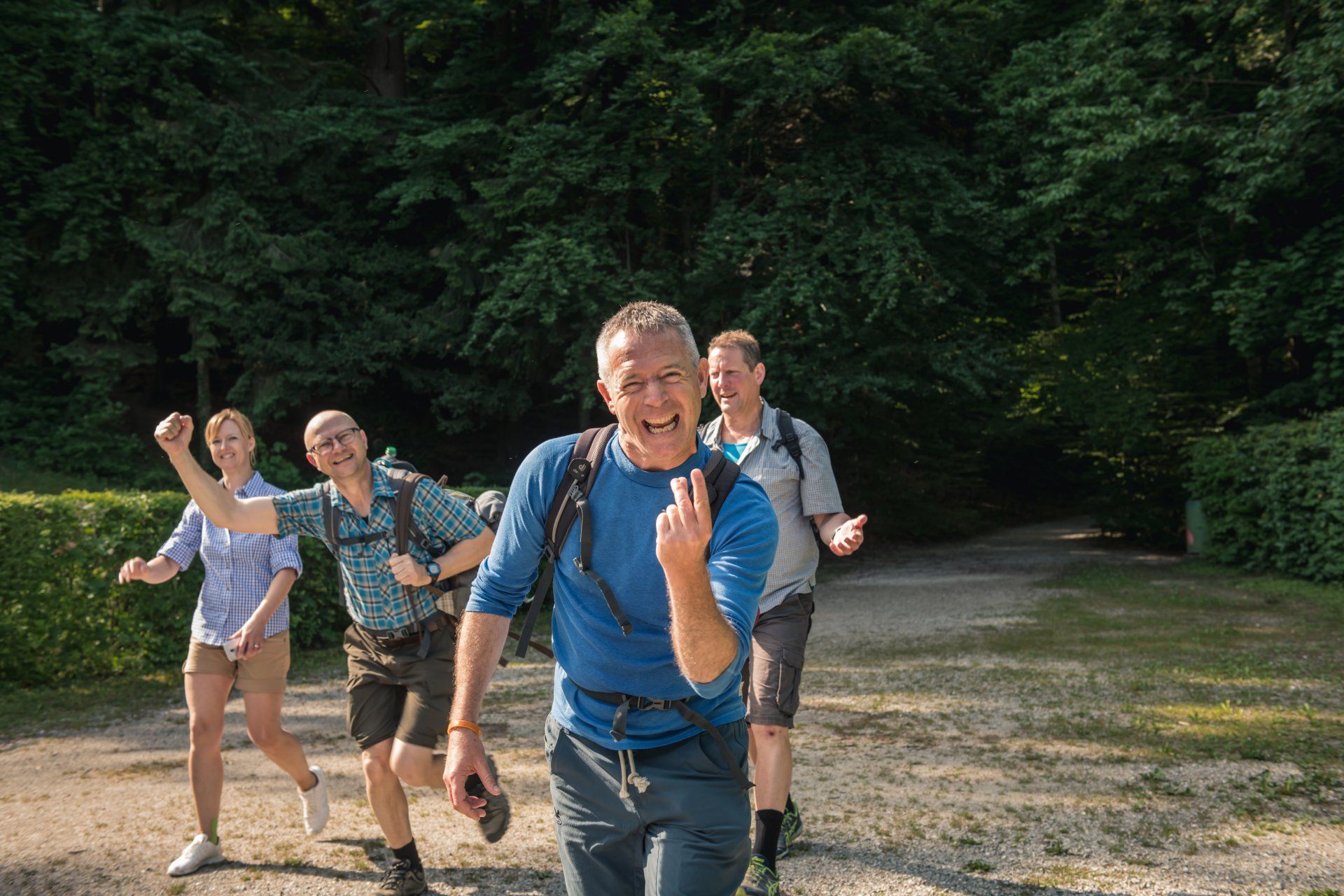 Eine Gruppe lächelnder Wanderer geht auf einem Pfad in einen Wald; einer macht das Peace-Zeichen.