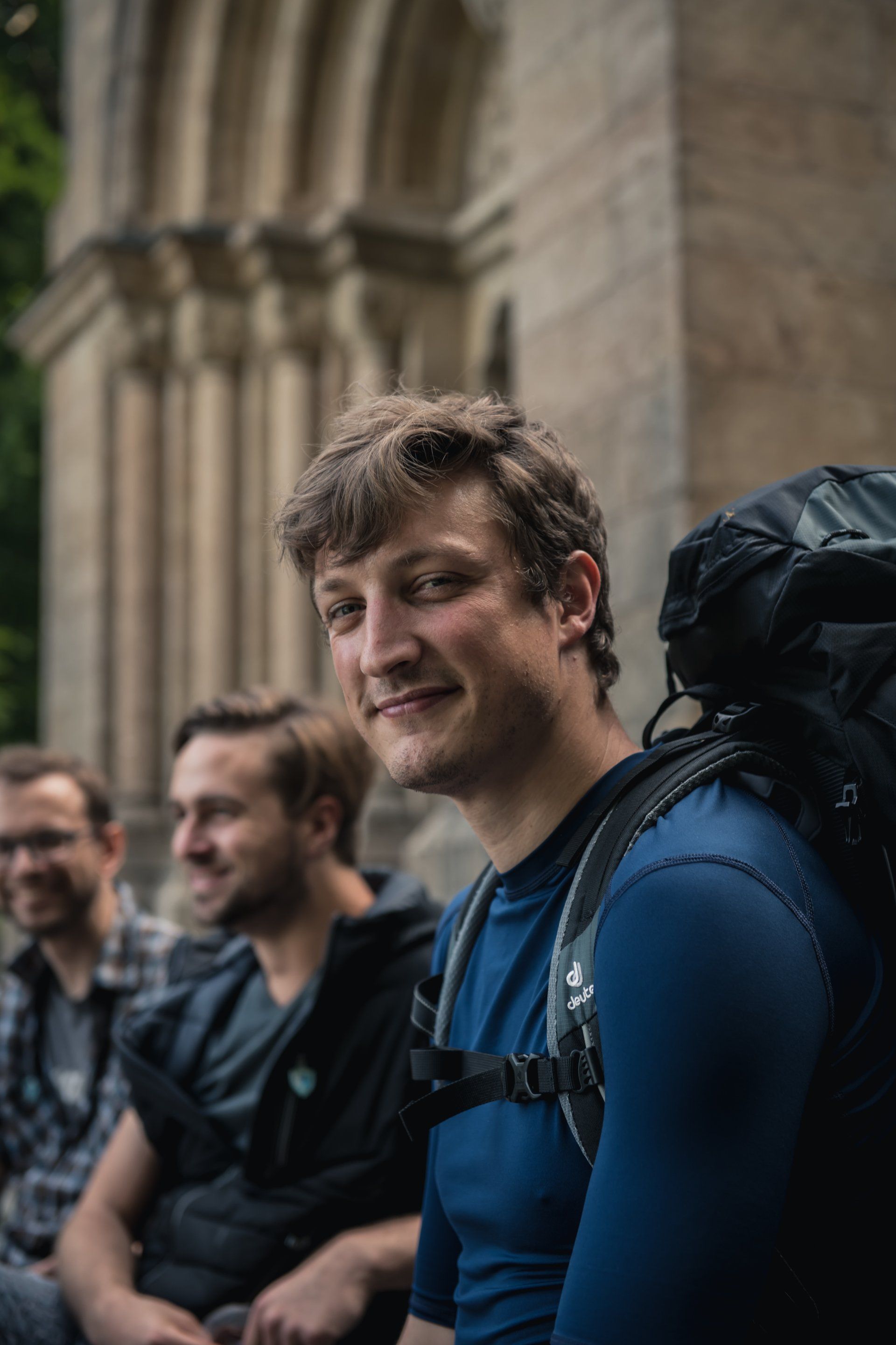 Lächelnder Mann mit Rucksack, posiert mit Freunden vor einem Steingebäude.