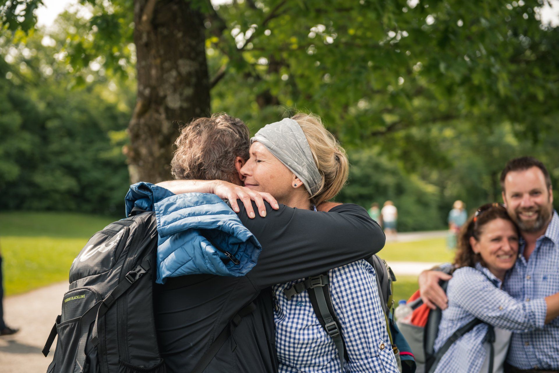 Zwei Paare umarmen sich im Freien; das Paar im Vordergrund umarmt sich unter einem Baum.
