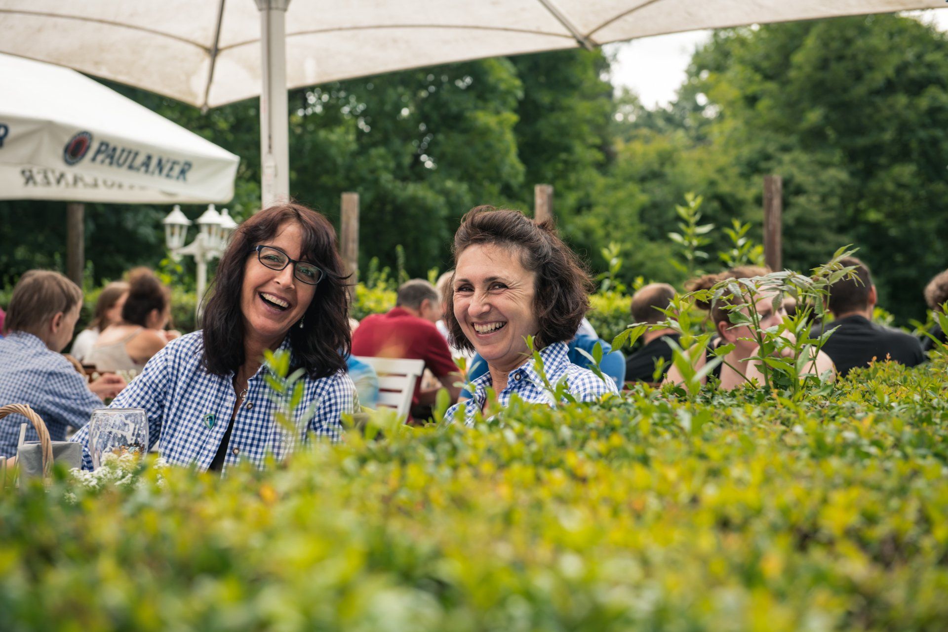 Zwei lachende Frauen, umrahmt von grünen Büschen, im Freien eines Restaurants.