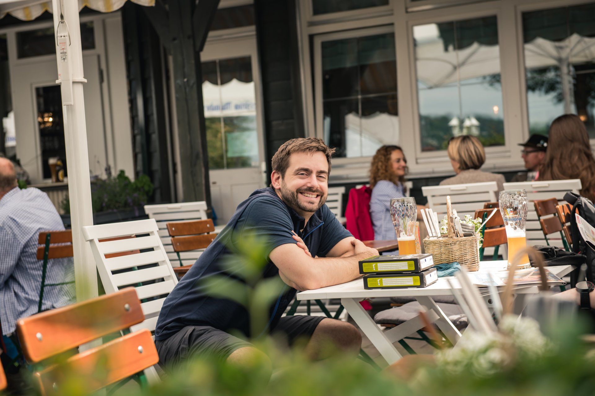 Mann lächelt in die Kamera. Er sitzt an einem Cafétisch im Freien. Andere Personen, Getränke und ein Gebäude sind zu sehen.