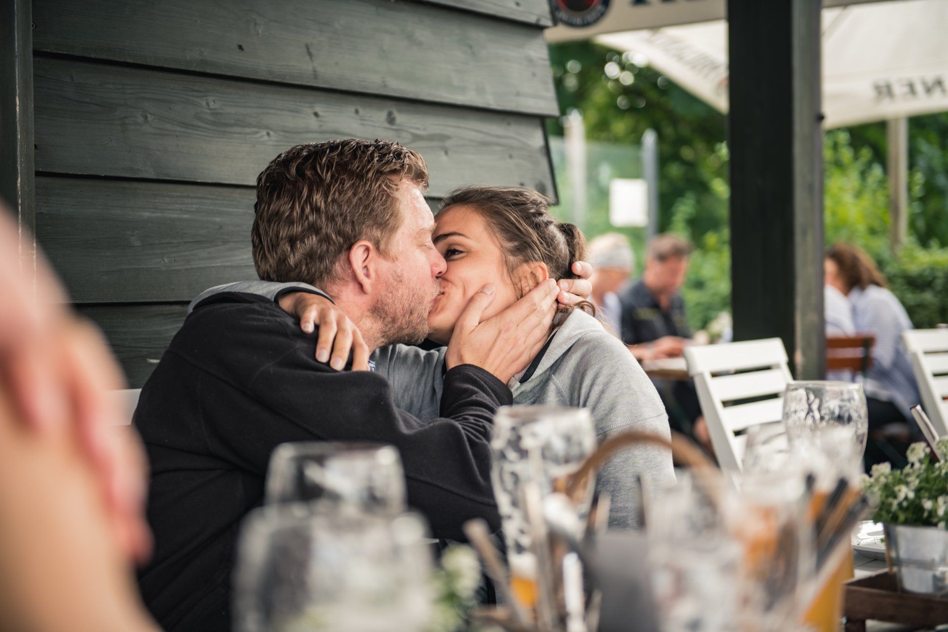 Paar küsst sich leidenschaftlich in einem Straßencafé; Holzhintergrund, andere Gäste sind im Hintergrund unscharf.