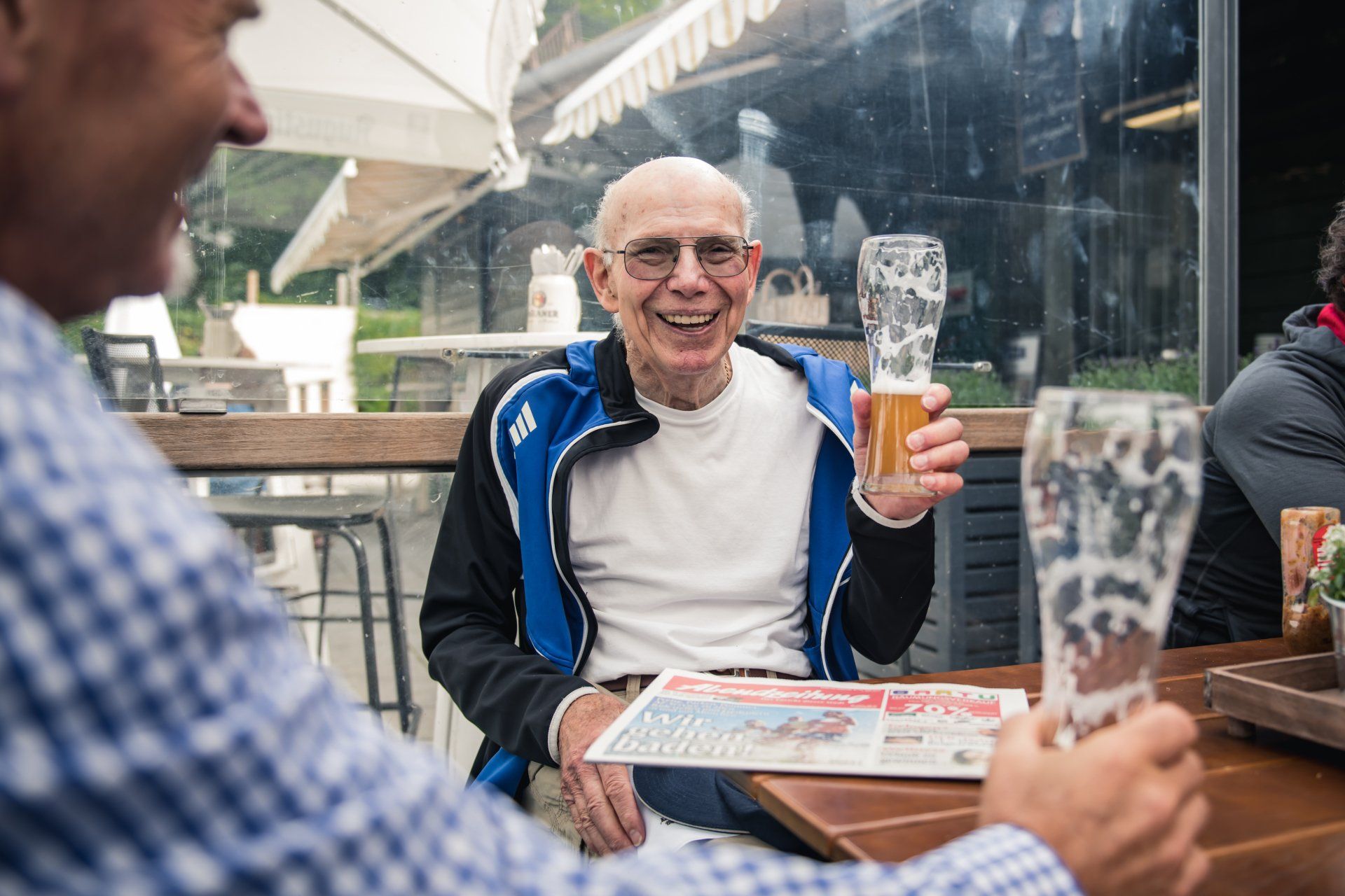 Zwei ältere Männer lächeln und stoßen mit Biergläsern an einem Tisch im Freien an.