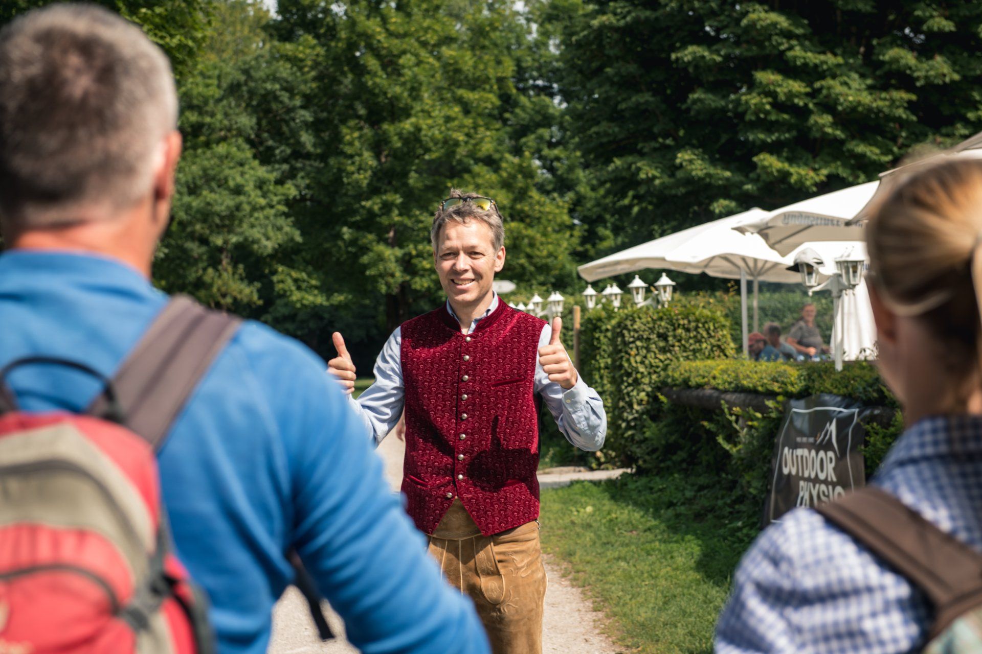 Mann in Weste und Lederhose zeigt Wanderern auf dem Weg in der Nähe eines Restaurants den Daumen hoch.
