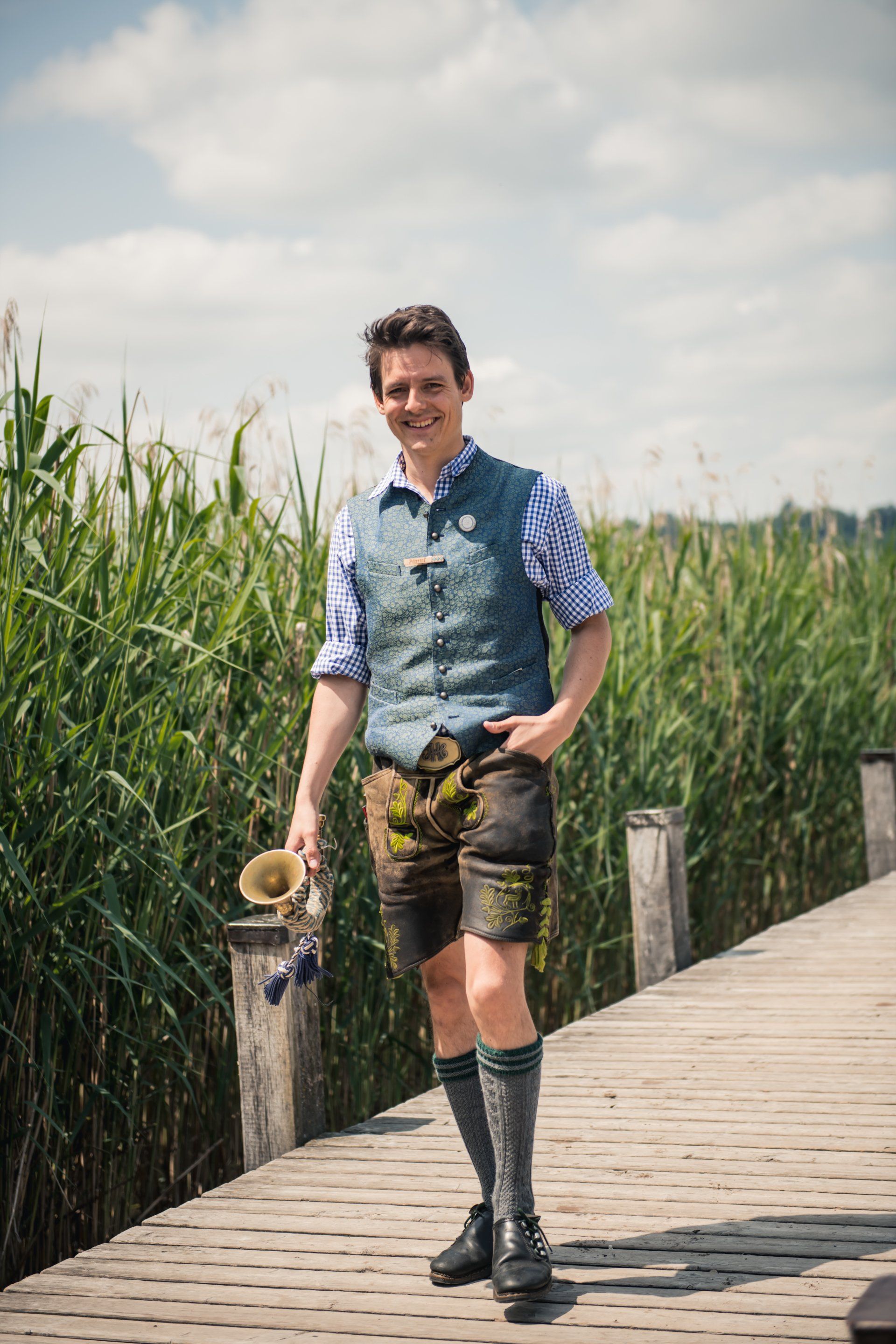 Mann in traditioneller bayerischer Kleidung hält eine Trompete auf einem Holzsteg in der Nähe von Schilf.