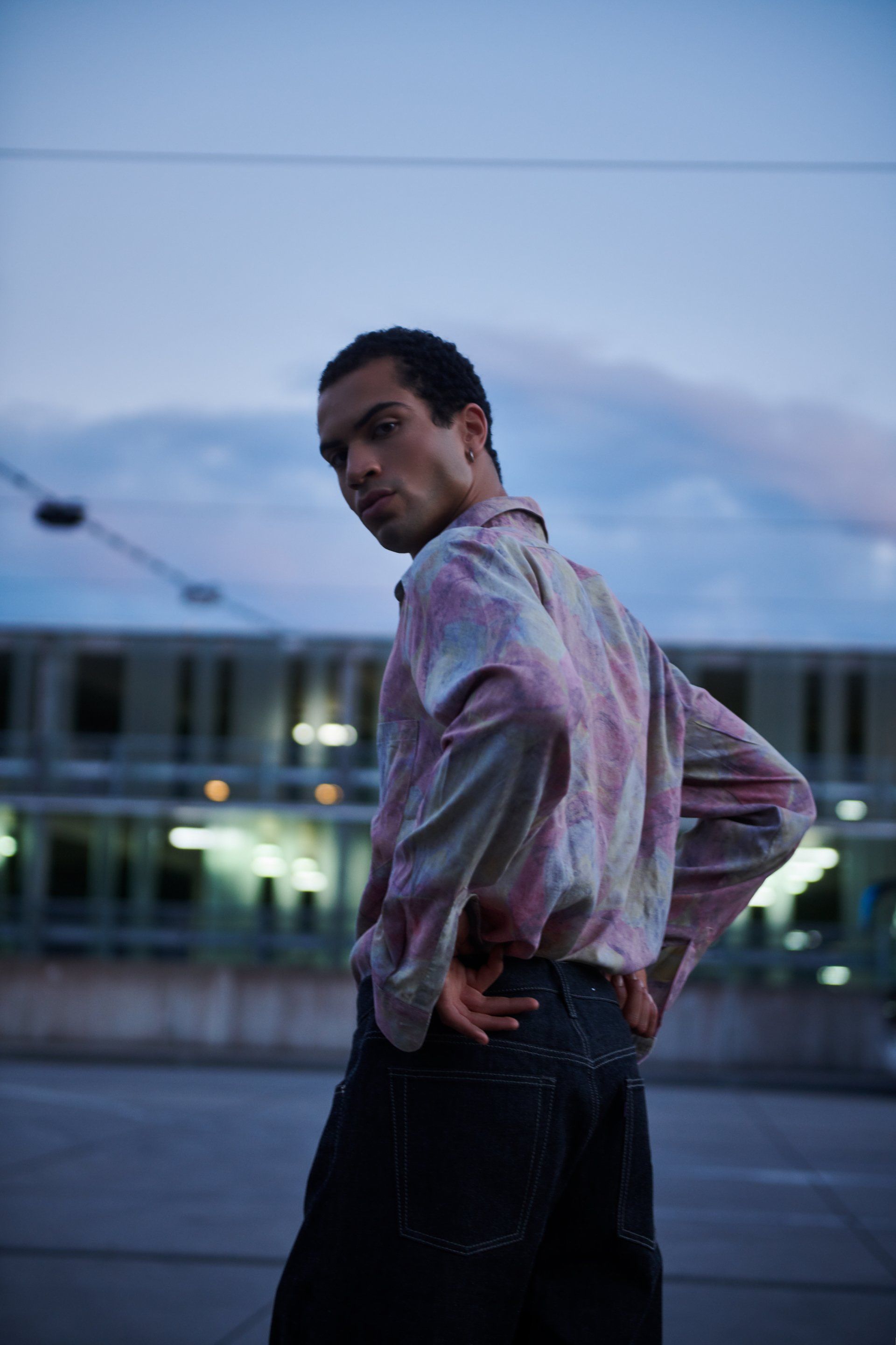 Mann in gemustertem Hemd und dunkler Jeans, Hände in die Hüften gestemmt, Blick über die Schulter; städtische Umgebung.