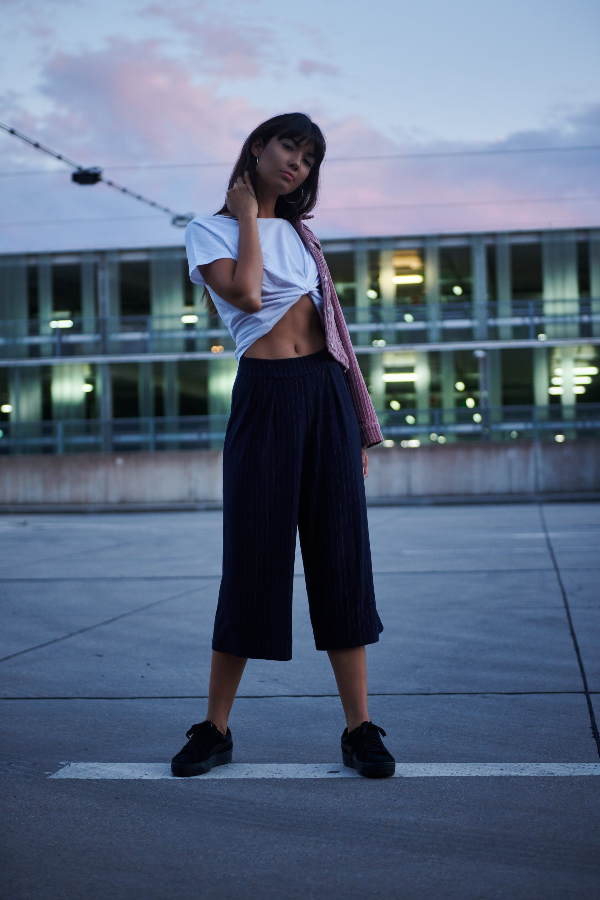 Frau in lässiger Kleidung auf dem Dach, blickt in die Kamera, mit Gebäude und Abendhimmel im Hintergrund.