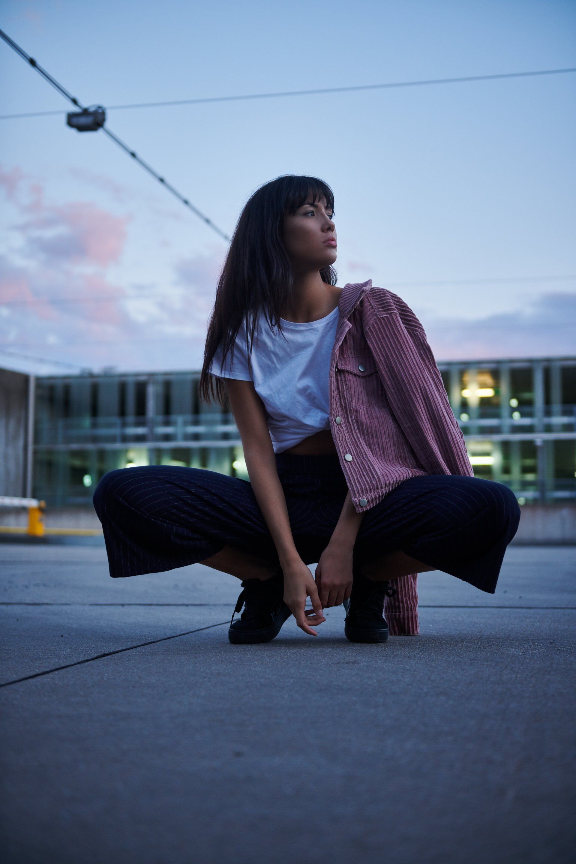 Frau in weißem T-Shirt, schwarzer Hose und rosa Jacke hockt auf einem Dach und blickt in den Abendhimmel.