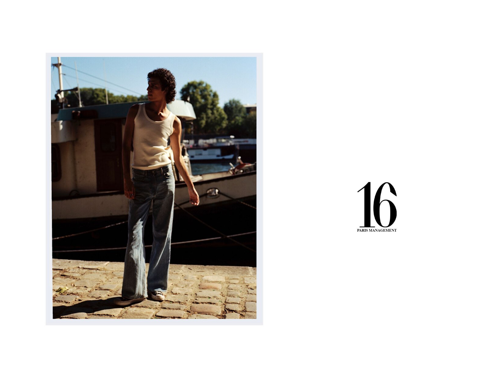 Mann in Tanktop und weiten Jeans steht neben einem Boot, sonnige Szene im Freien.