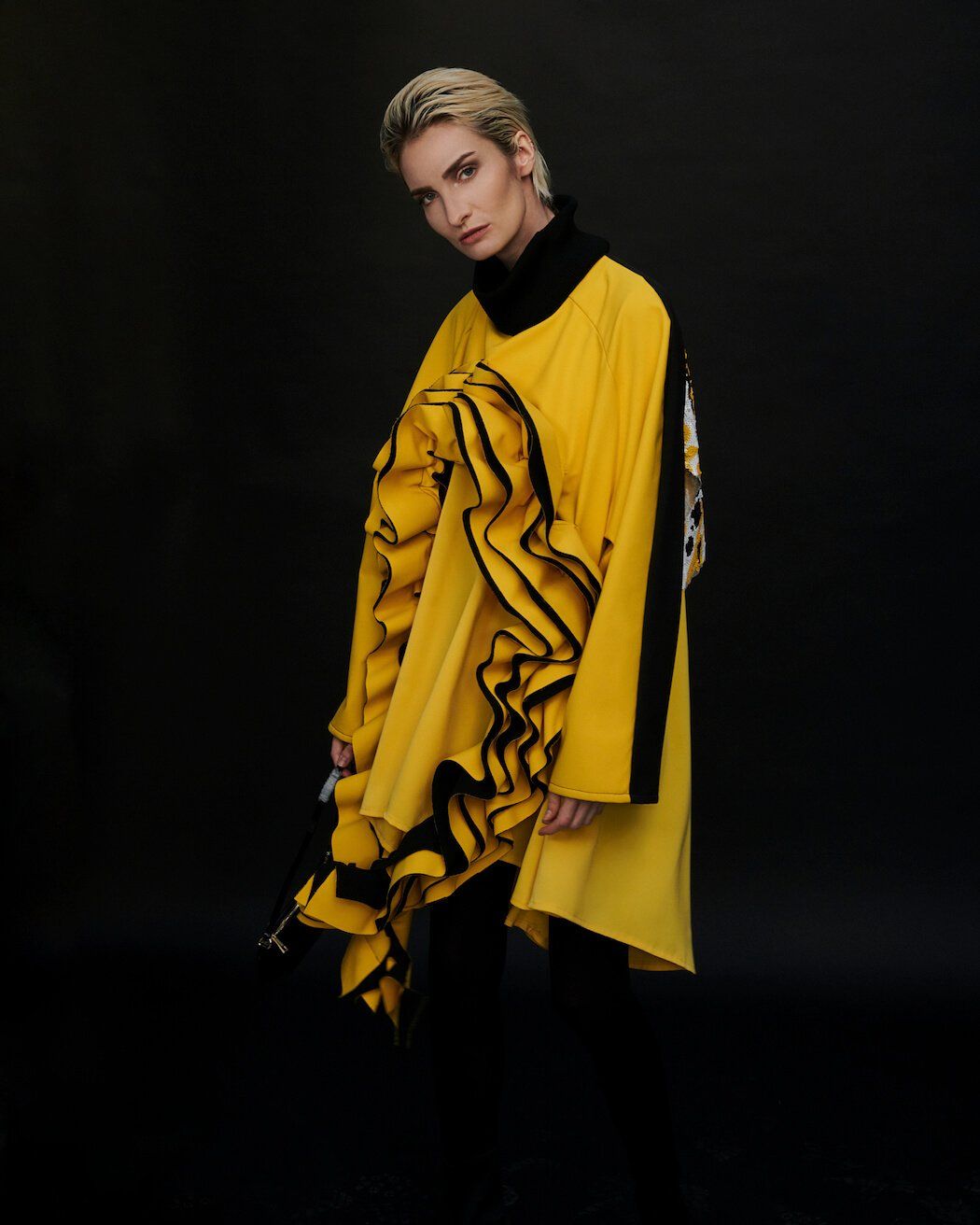 Frau in gelbem Rüschentop, schwarzem Rollkragenpullover und kurzen blonden Haaren vor schwarzem Hintergrund.