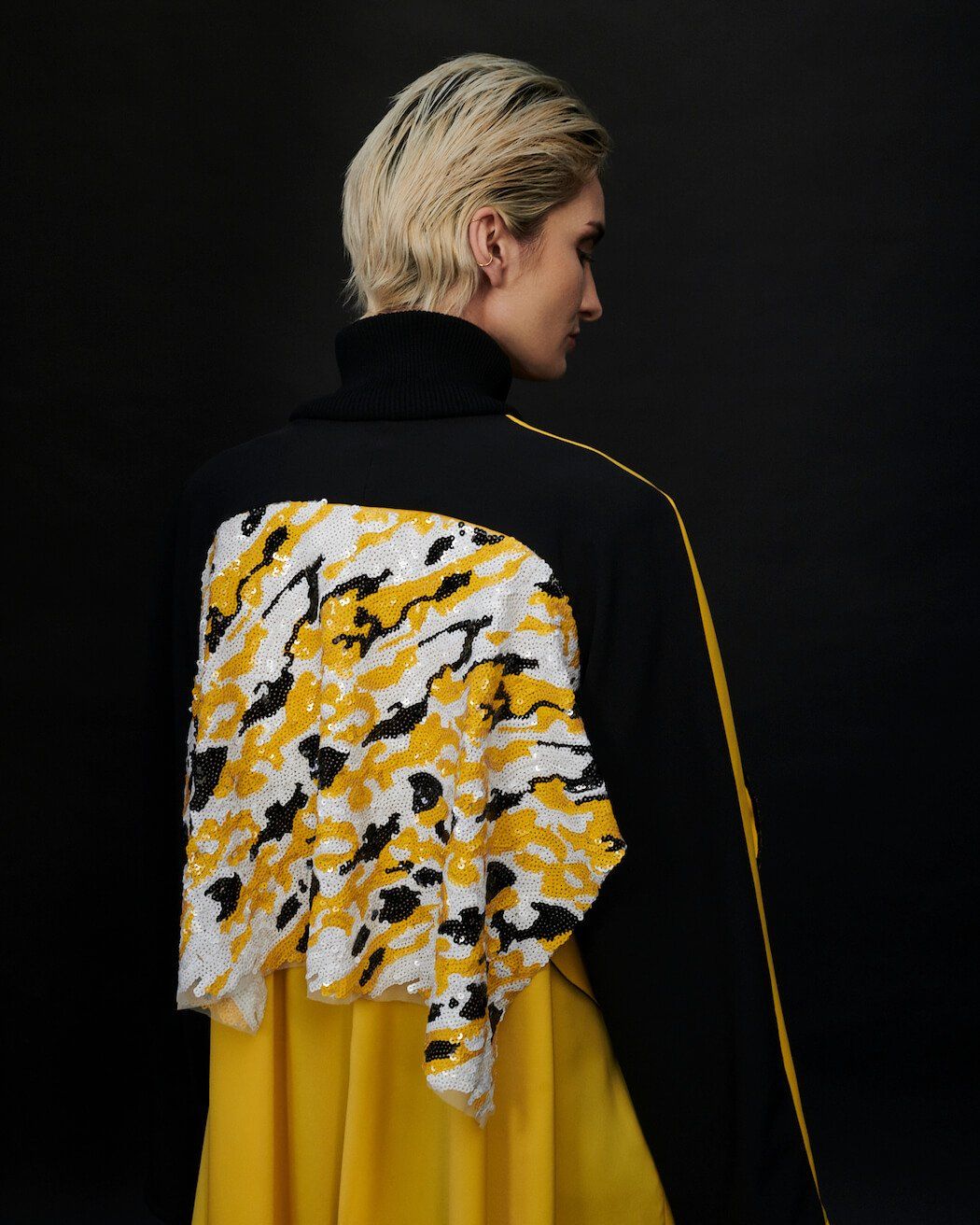 Person in schwarz-gelber Jacke, Rücken zur Kamera, blondes Haar, gelbes Muster auf dem Rücken, schwarzer Hintergrund.