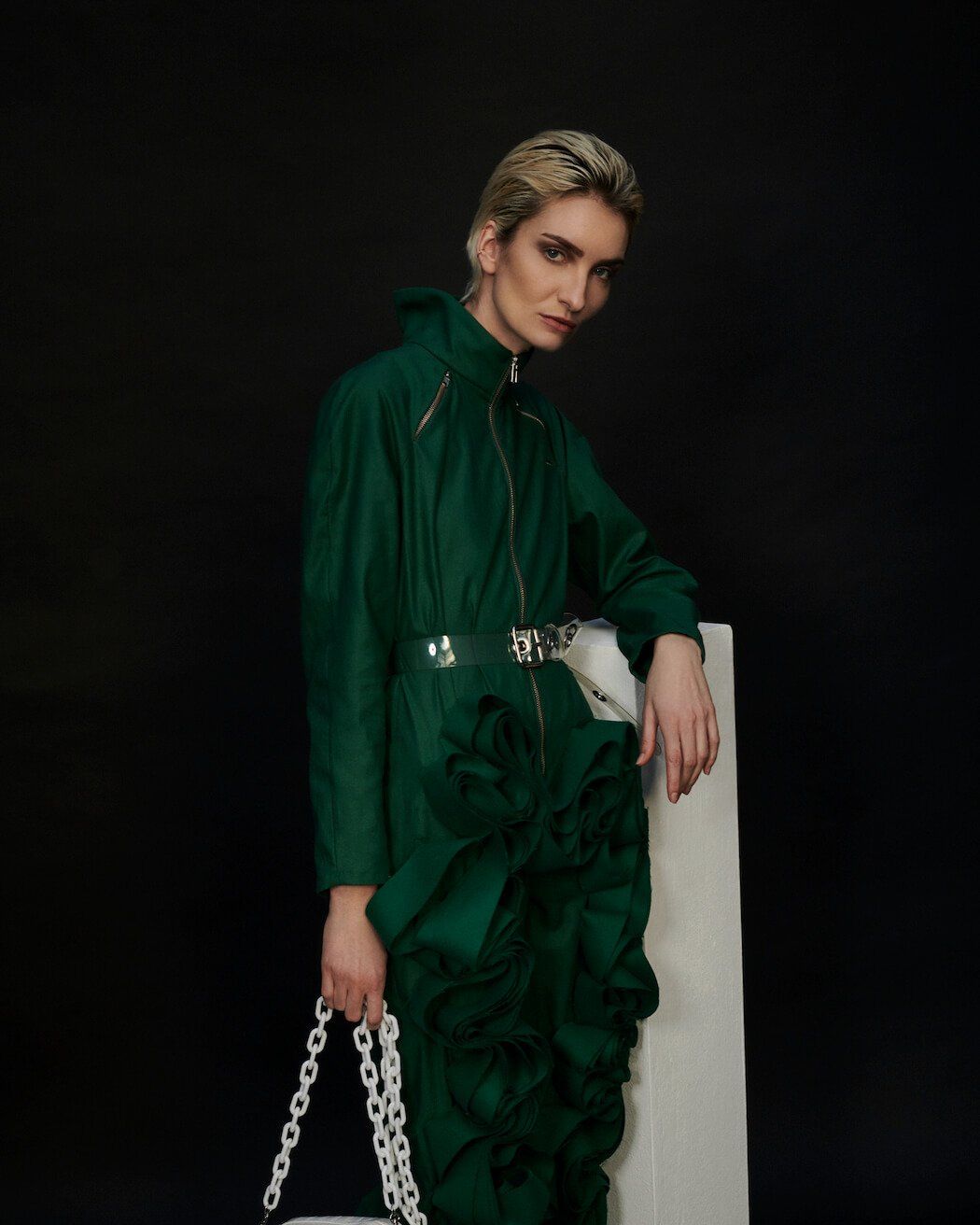 Person mit kurzen blonden Haaren, in grünem Overall mit weißer Kettentasche, posiert vor schwarzem Hintergrund.