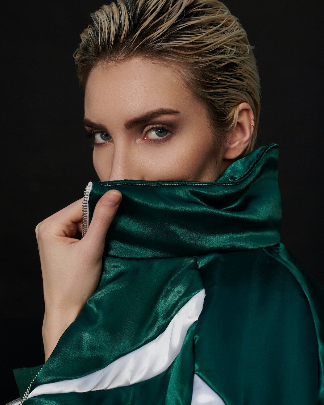 Frau mit kurzen blonden Haaren, grüner Jacke, über den Kragen blickend.