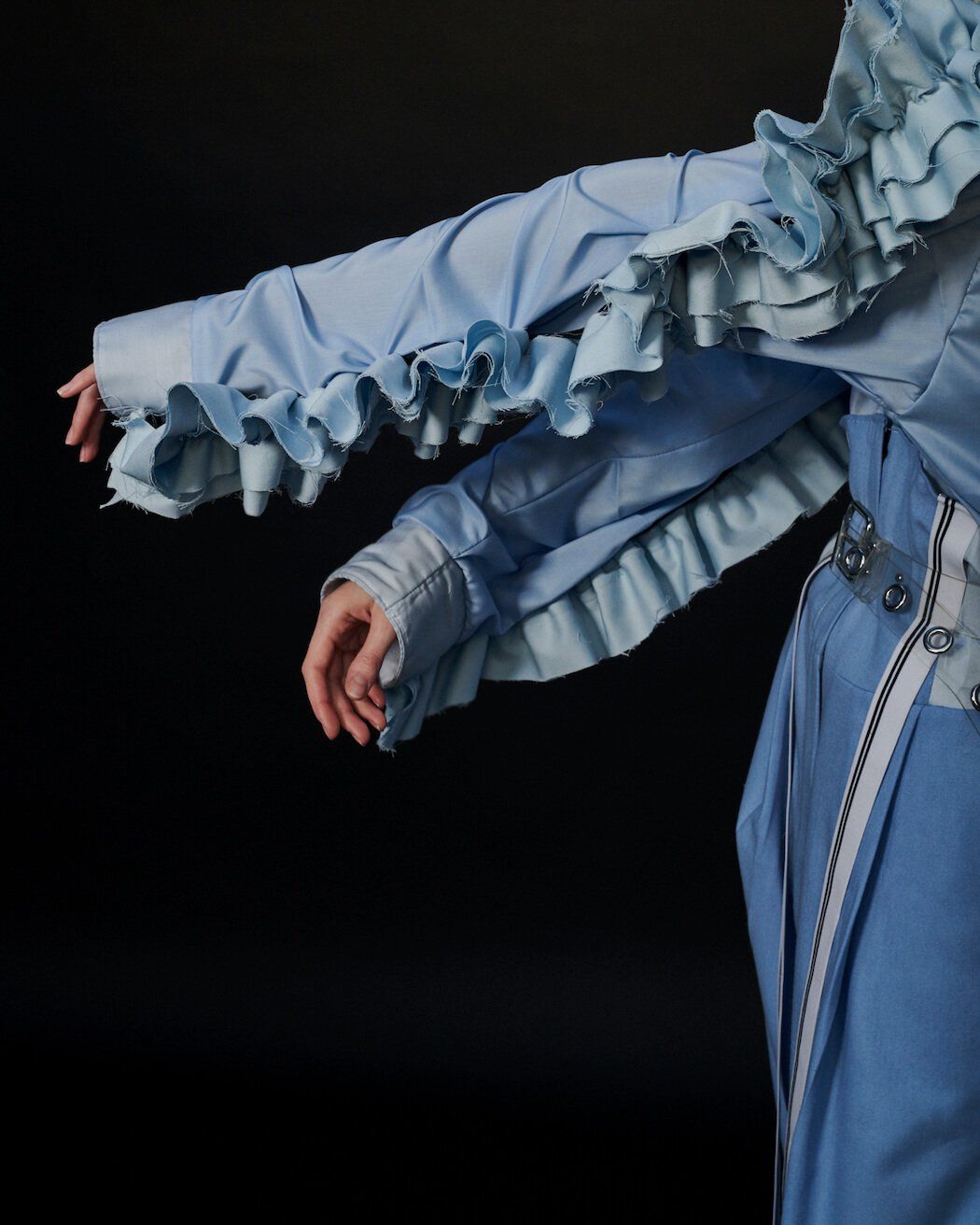 Arm einer Person in hellblauer Kleidung mit Rüschenärmeln. Dunkler Hintergrund.
