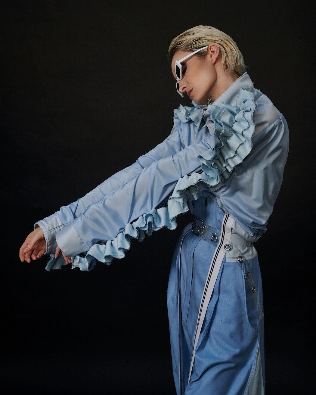 Person mit kurzen blonden Haaren, Jeans-Outfit mit Rüschen, weißer Sonnenbrille, ausgestreckten Armen.