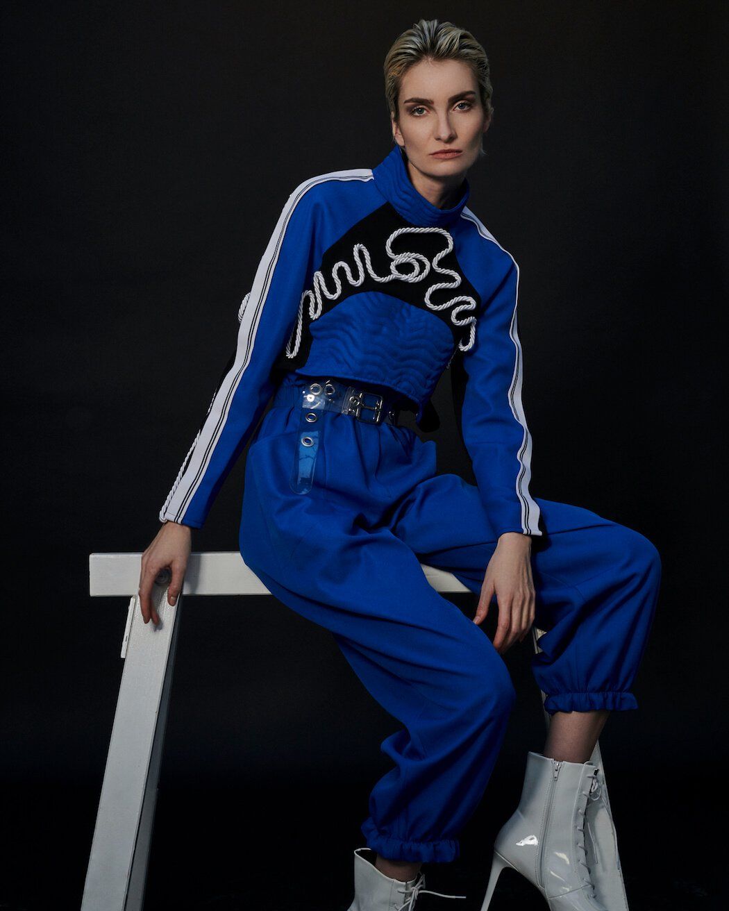 Frau im blauen Outfit, sitzt auf weißem Hocker. Schwarzer Hintergrund.