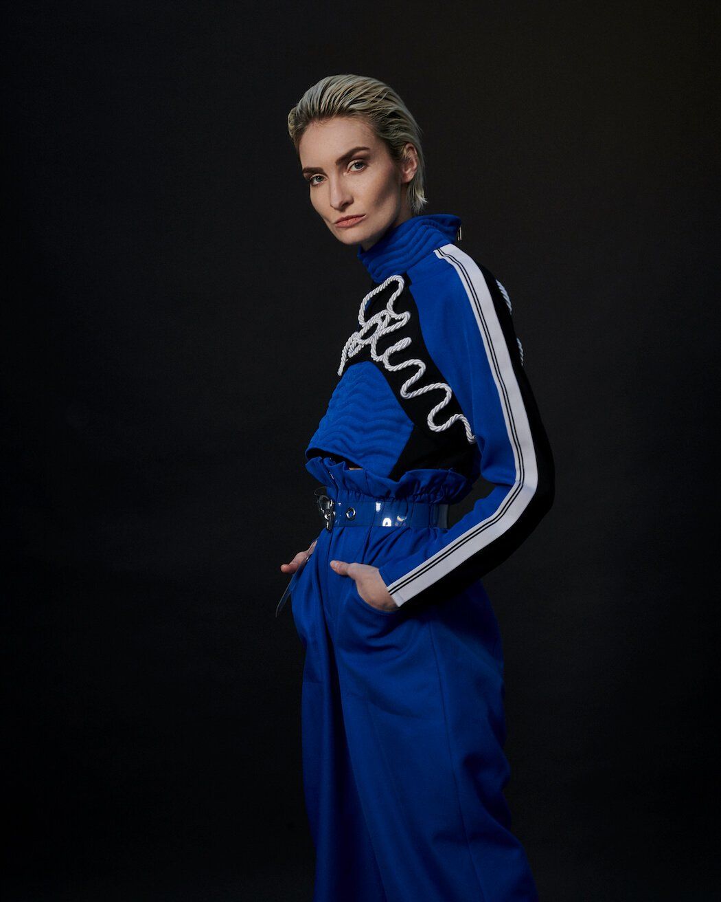 Frau im blauen Overall mit weißen und schwarzen Akzenten, Hände in den Taschen, blickt in die Kamera.