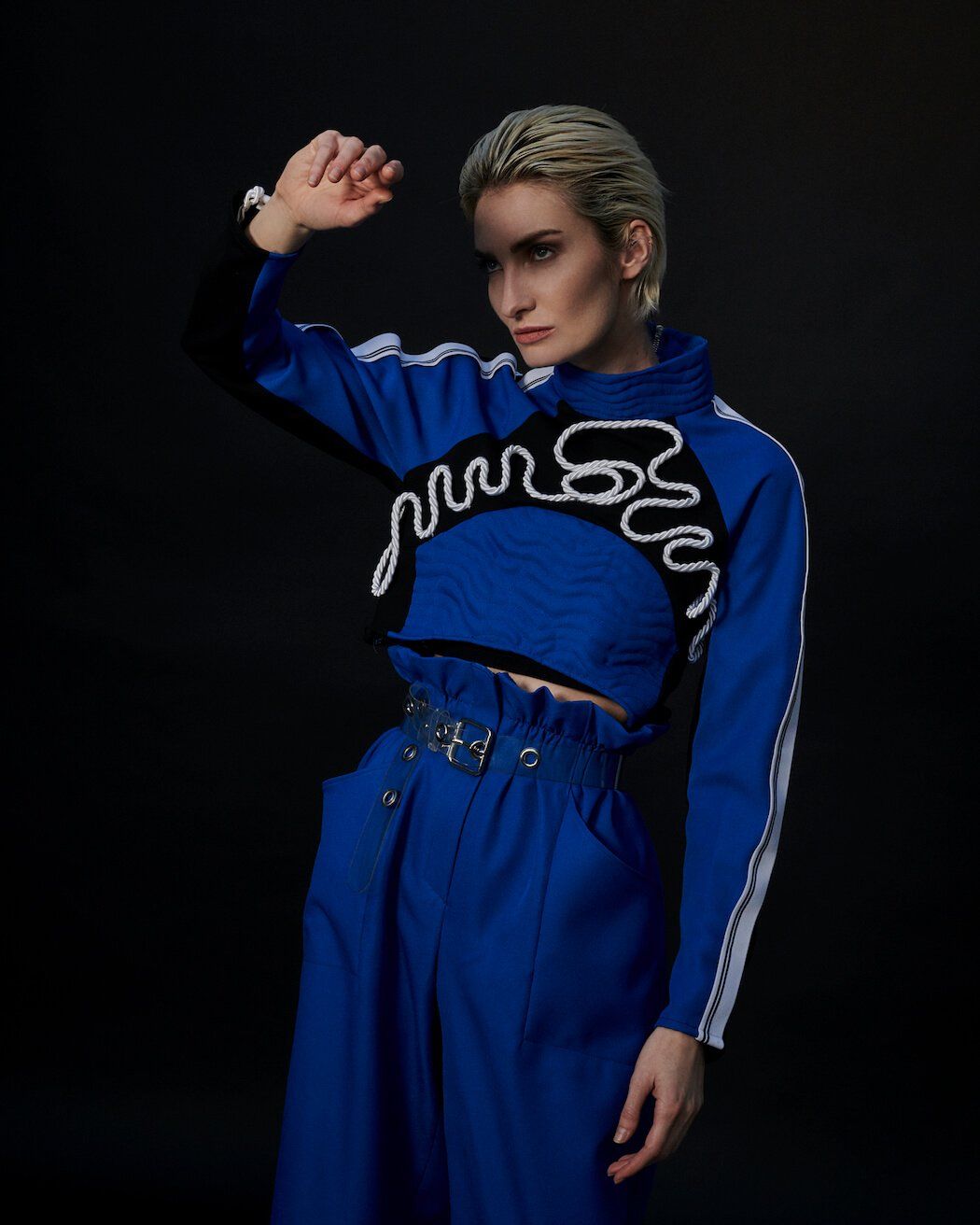 Frau in blauem bauchfreiem Top und Hose, Arm erhoben, blickt zur Seite vor dunklem Hintergrund.