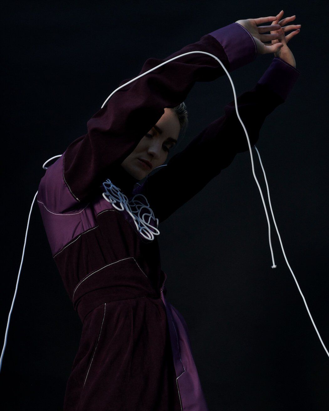 Frau in dunkelviolettem Mantel mit weißem Besatz, die Arme erhoben, vor schwarzem Hintergrund.