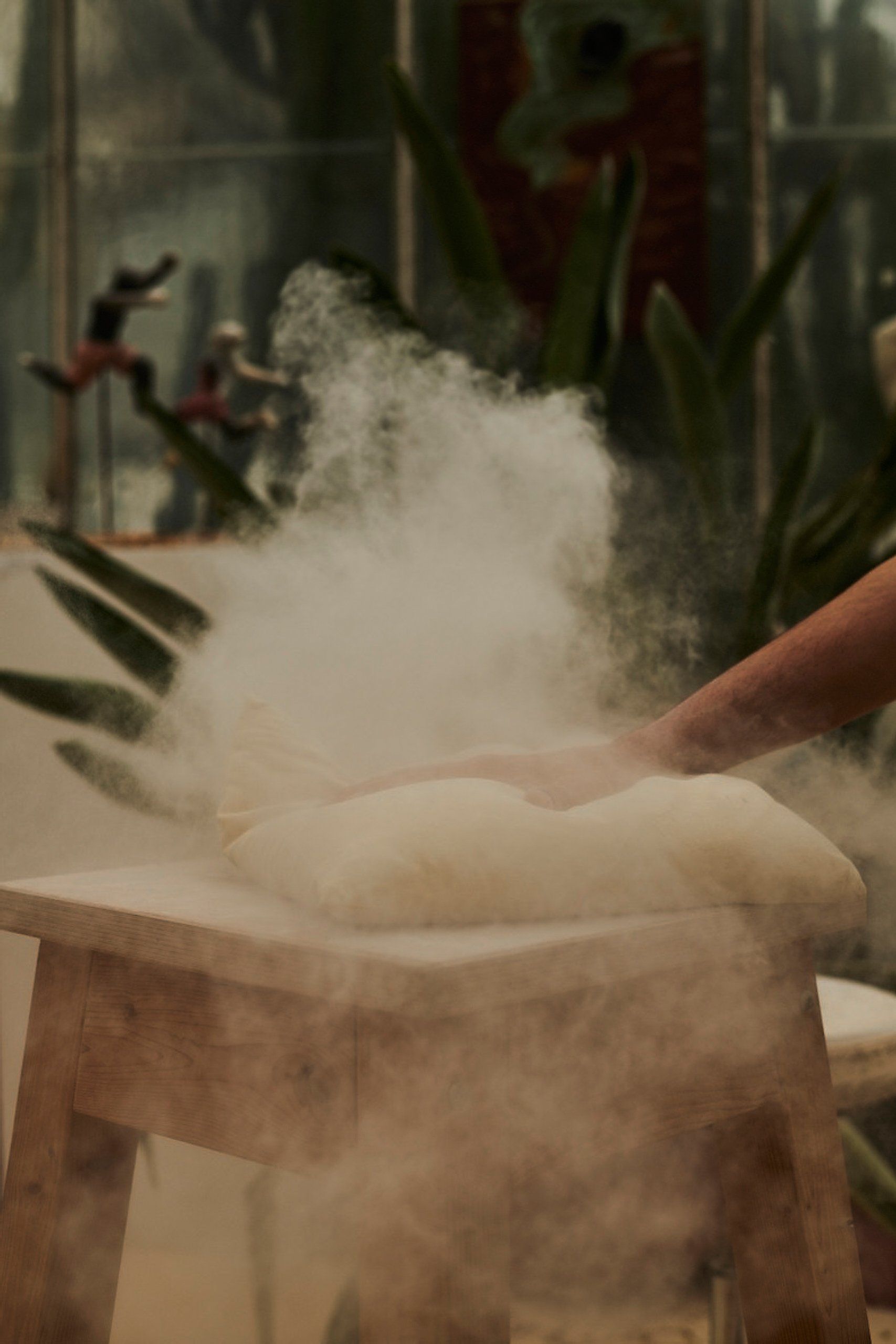 Die Hand einer Person drückt auf mit Mehl bestäubten Teig und erzeugt so eine Wolke aus weißem Staub auf einem Holztisch.
