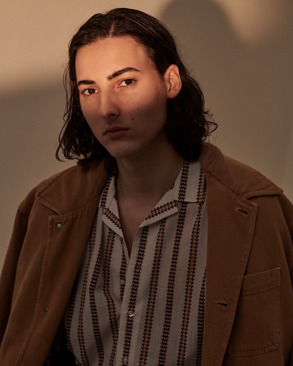 Mann mit langen dunklen Haaren in Cordjacke und gestreiftem Hemd, blickt in die Kamera.