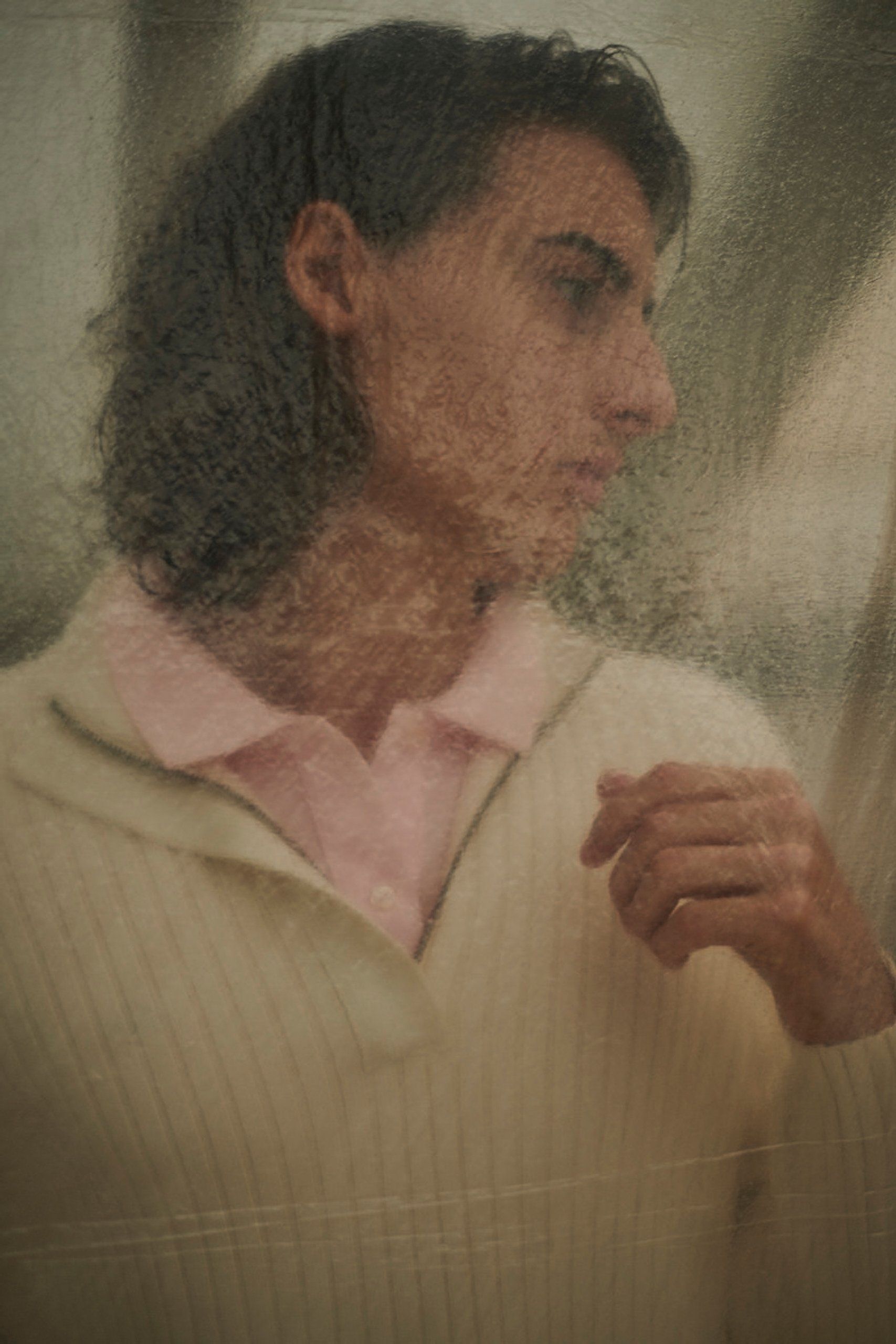Mann mit langen Haaren, hellem Pullover und rosa Hemd mit Kragen, blickt nachdenklich durch einen hauchdünnen Bildschirm.