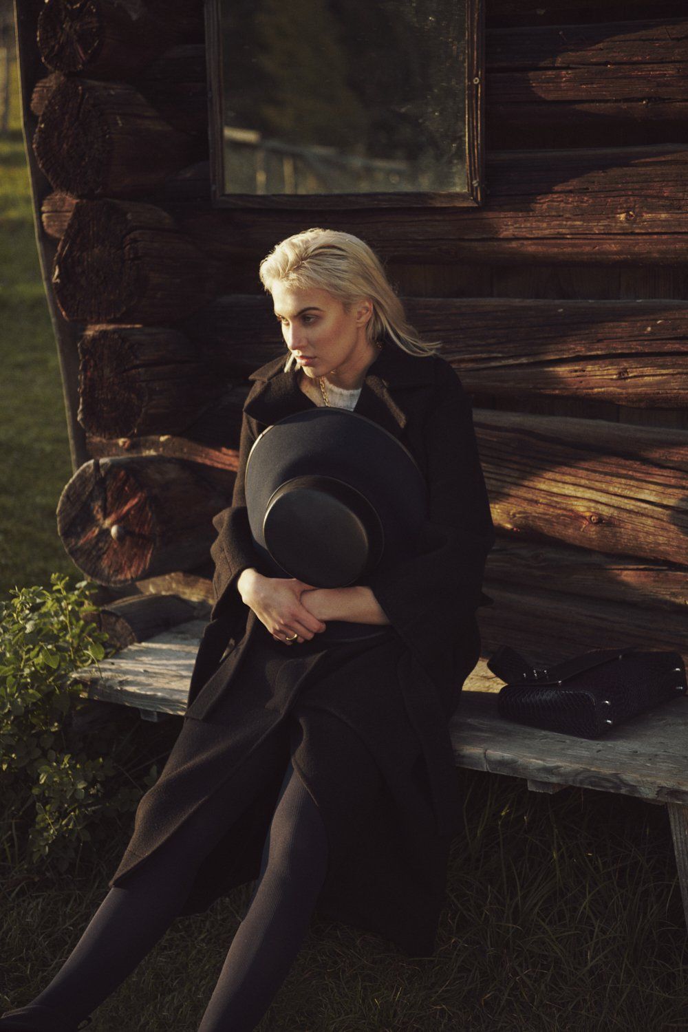 Frau im dunklen Mantel sitzt neben einem Holzgebäude und hält einen schwarzen Hut. 