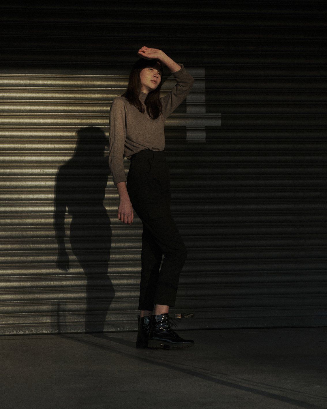 Frau, die ihre Augen abschirmt und vor einer Wellblechwand steht. Sie trägt ein braunes Hemd und eine schwarze Hose.