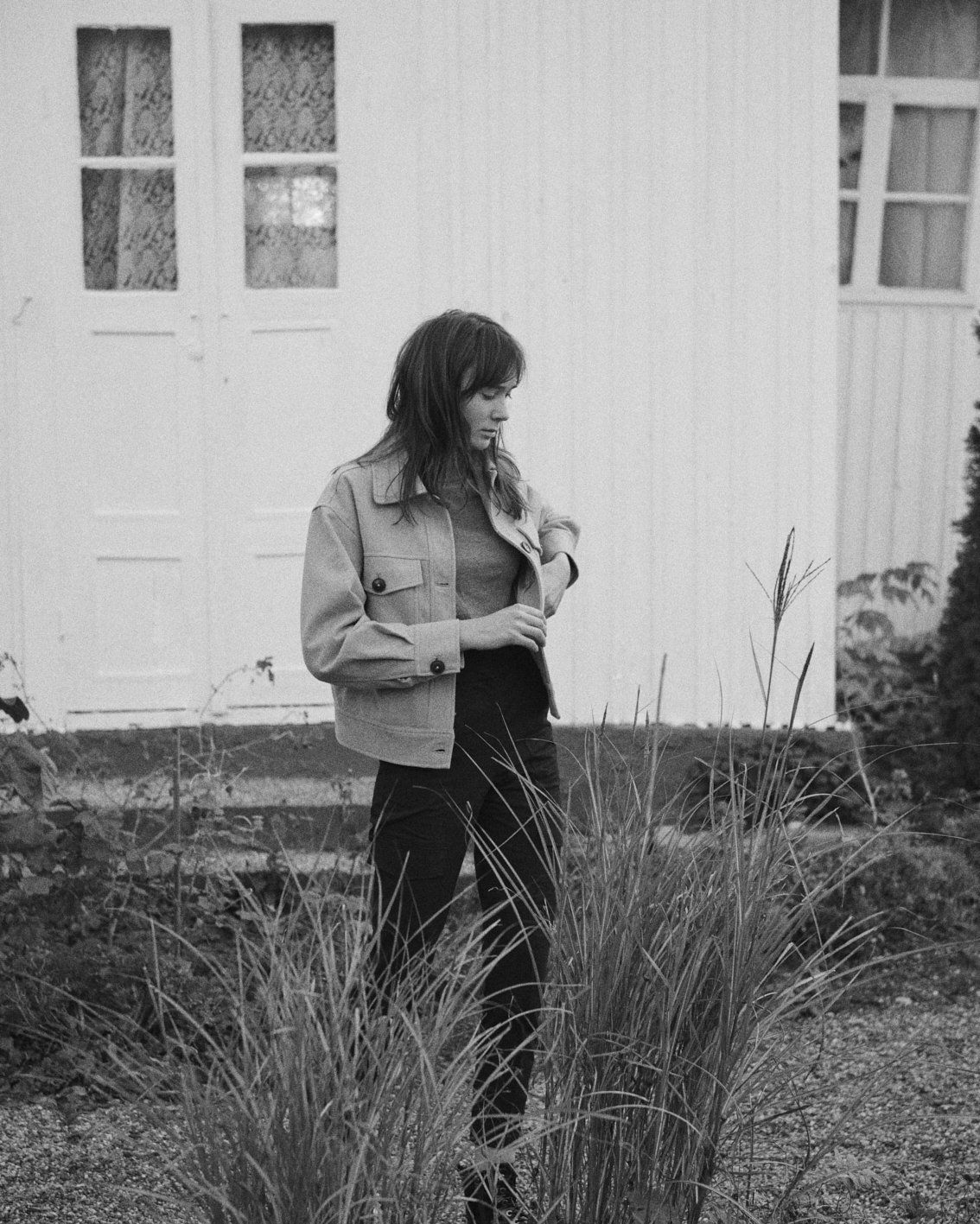 Frau in Jacke und Hose steht neben hohem Gras vor einem weißen Gebäude.
