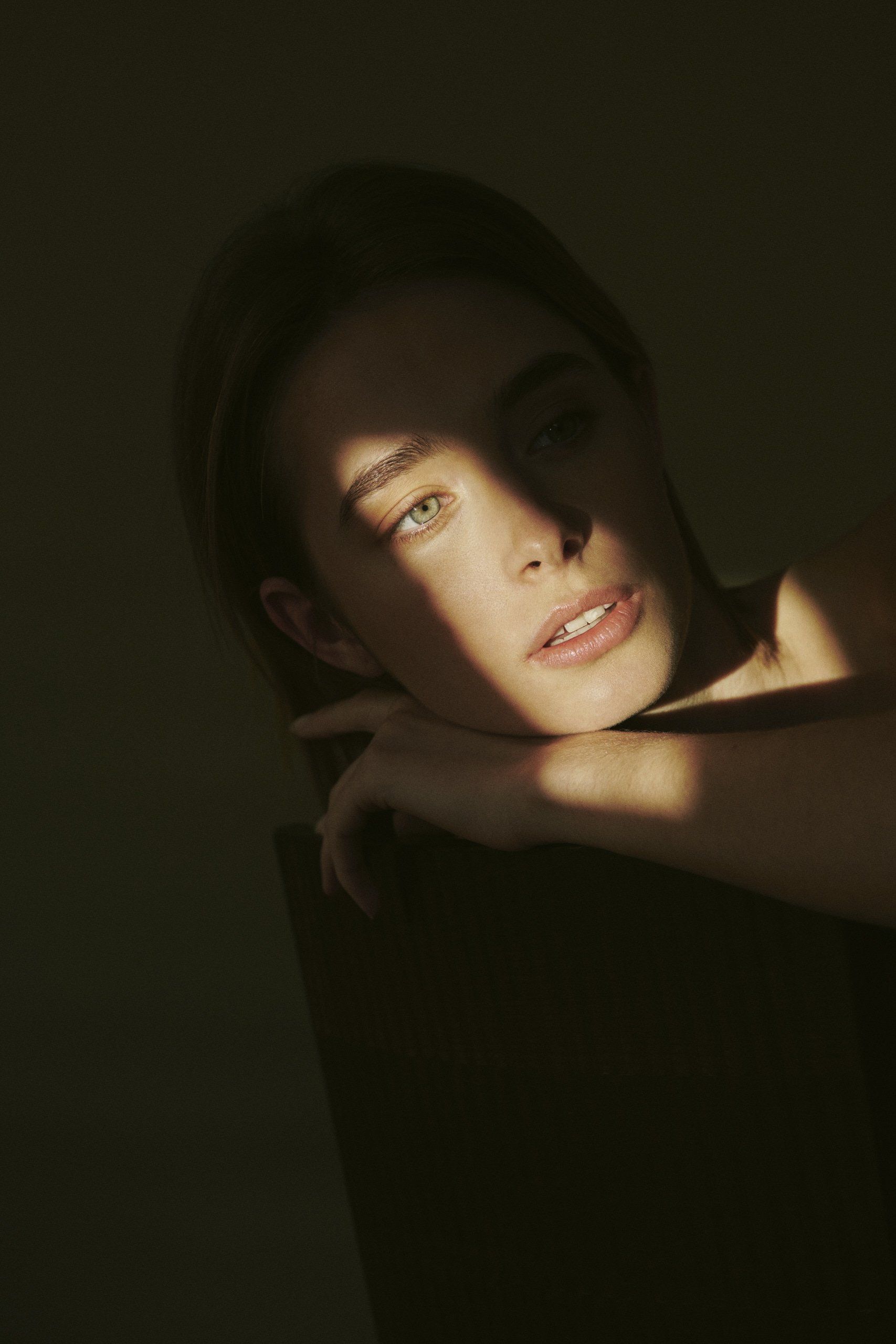 Frau mit sonnenbeschienenem Gesicht, das Kinn auf einem Stuhl ruhend, der Blick leicht vom Bild abgewandt.