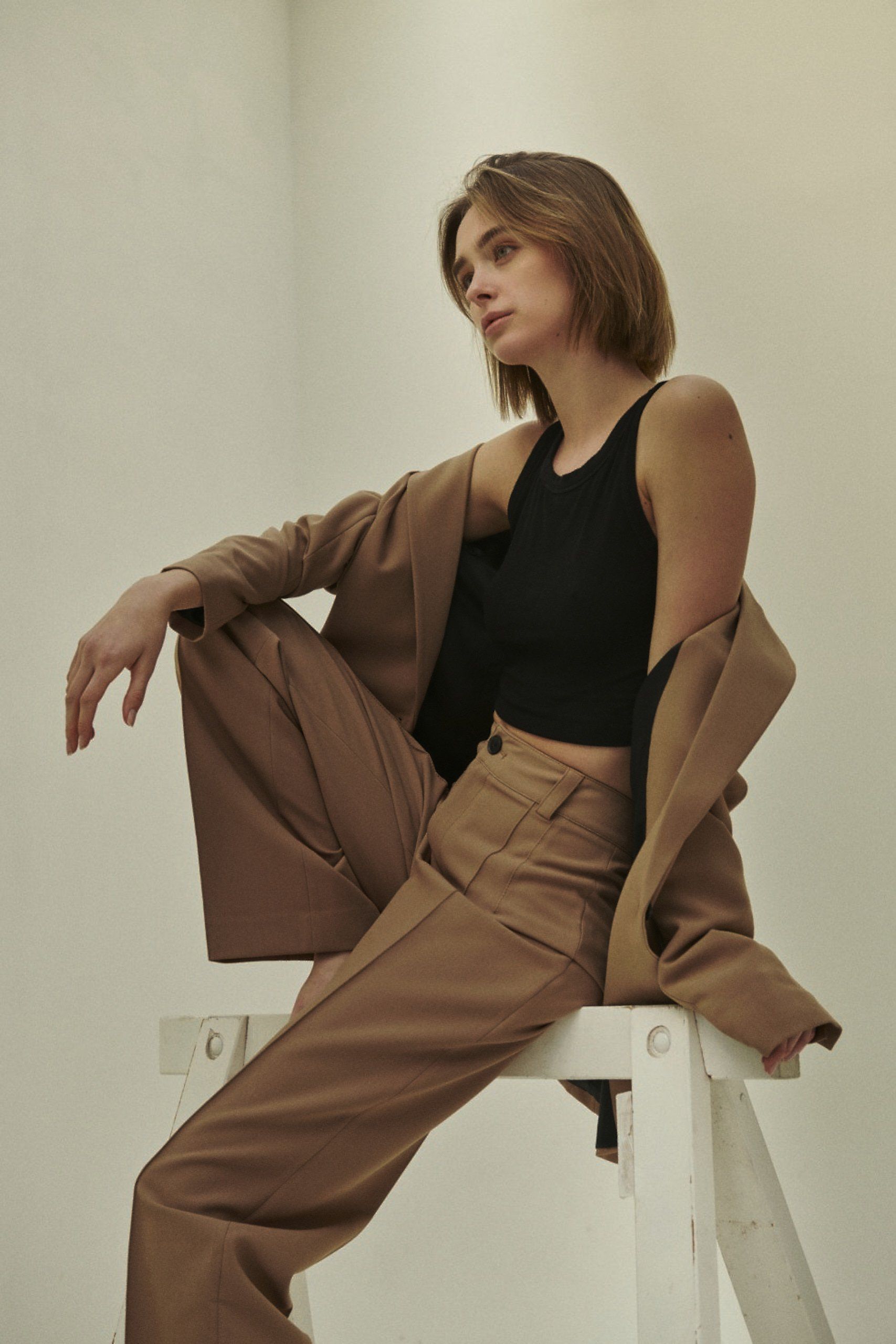 Frau in braunem Anzug und schwarzem Oberteil sitzt auf einem weißen Hocker und blickt zur Seite.