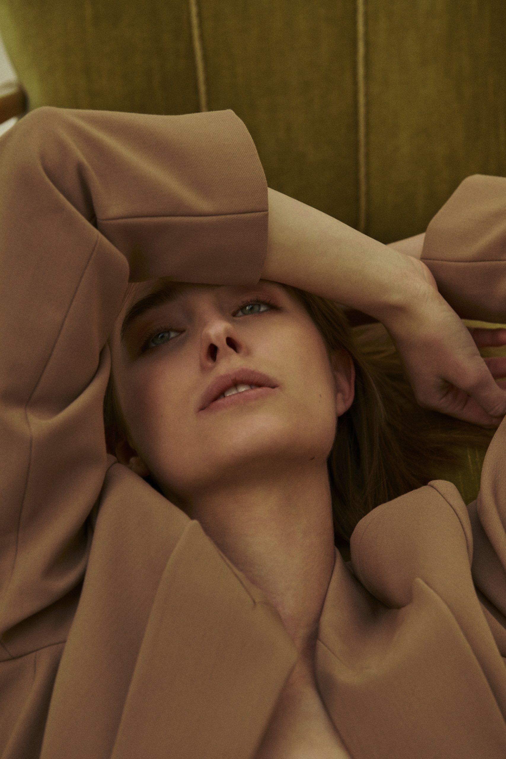 Blonde Frau im hellbraunen Mantel, die Augen nach oben gerichtet, ruht auf einem grünen Sofa, die Hand auf der Stirn.