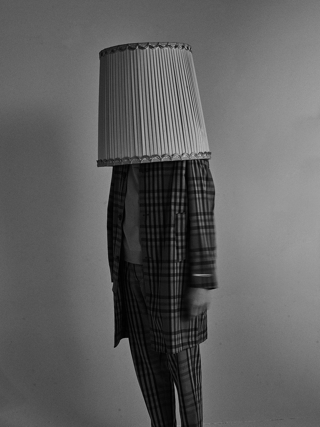 Person mit einem Lampenschirm als Kopf, kariertem Pyjama, steht vor einer neutralen Wand.