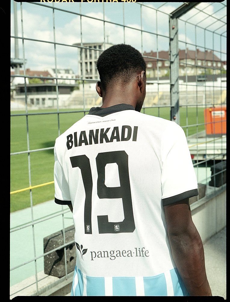 Mann in weißem Trikot mit „BIANKADI“ und „19“ auf dem Rücken, steht neben einem Zaun mit Blick auf ein Stadion.