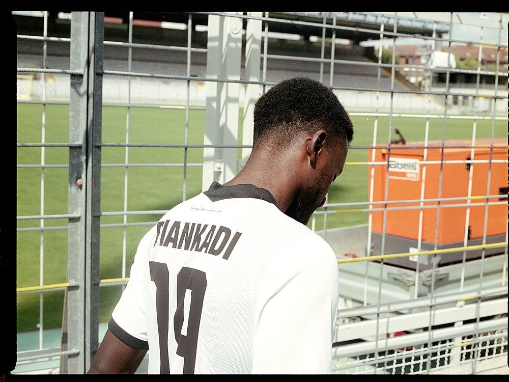 Mann im Fußballtrikot „TIANKADI 19“ lehnt am Zaun des Stadions und blickt nach unten.