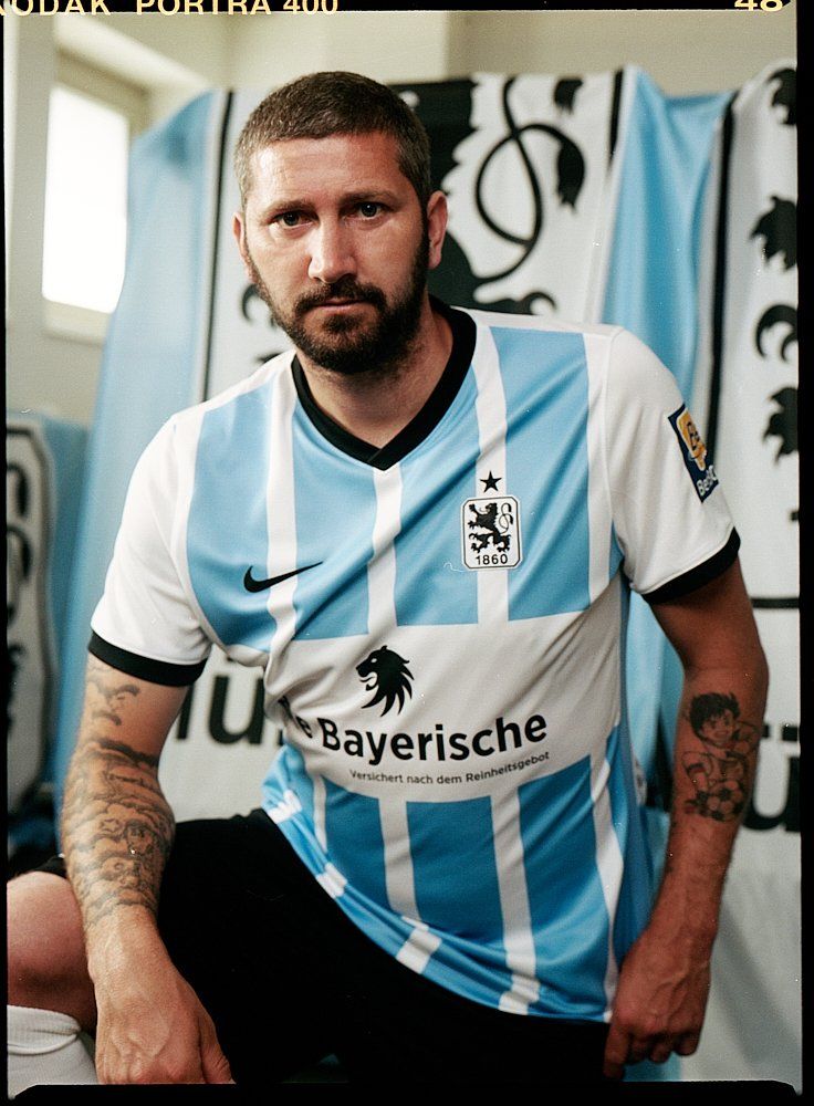 Mann in blau-weißem Fußballtrikot, mit Tattoos auf dem Arm, steht vor der Mannschaftsflagge.