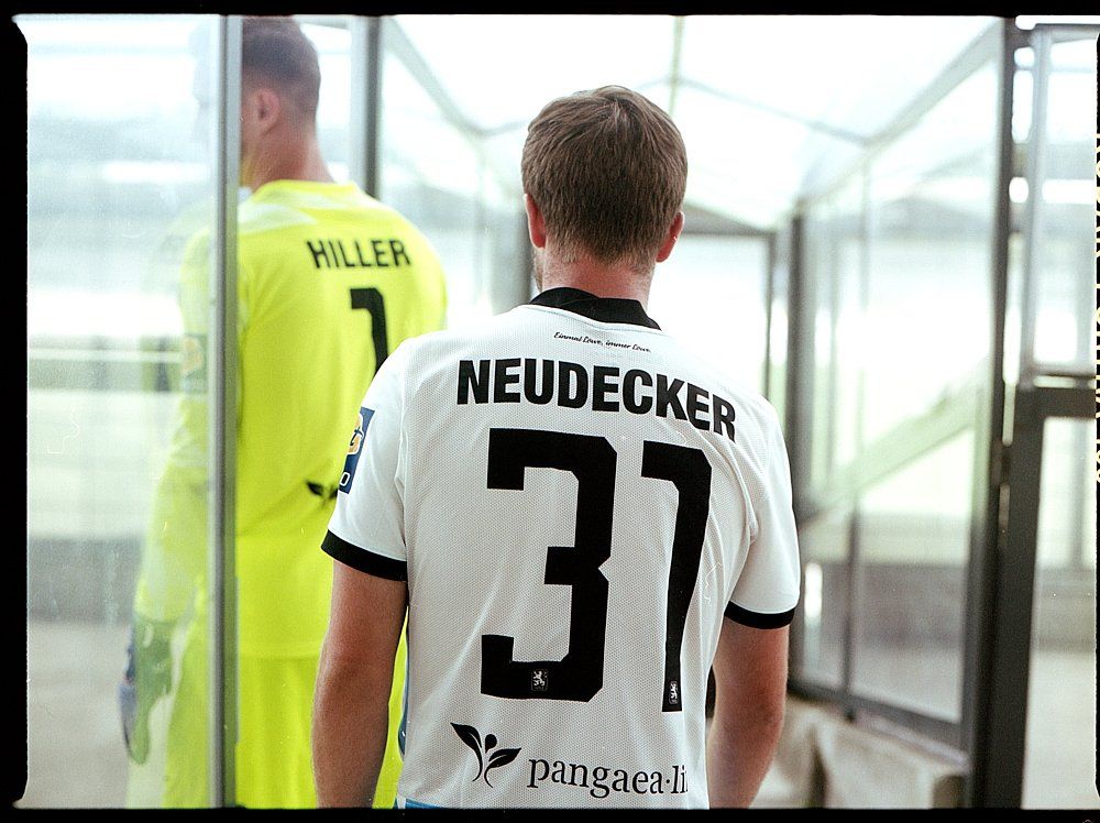Fußballspieler im Tunnel; Trikot mit „NEUDECKER 37“ auf dem Rücken, Torwart in Gelb dahinter, „HILLER 1“.