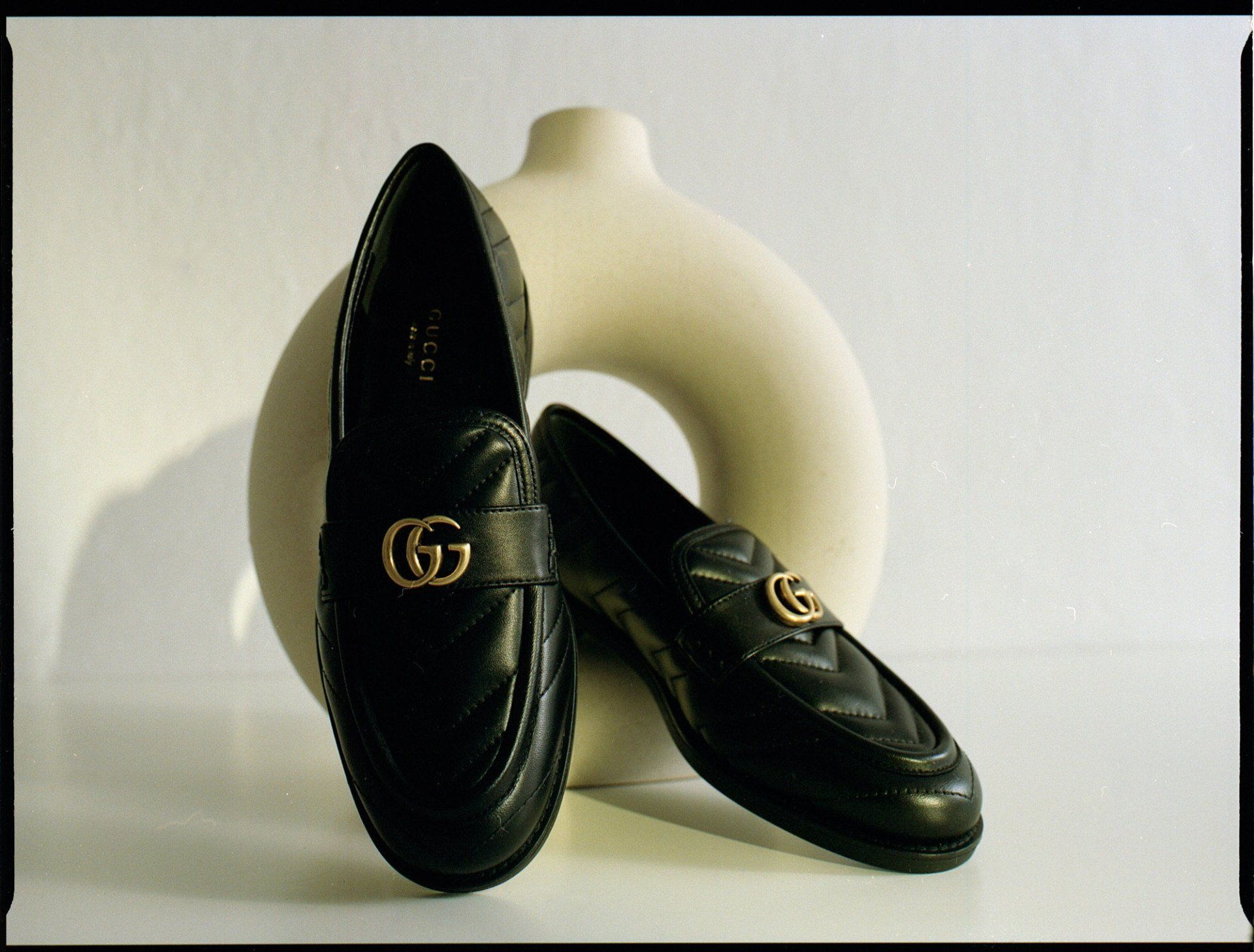 Schwarze Gucci-Loafer mit goldenem Logo und beiger Vase auf weißem Untergrund.