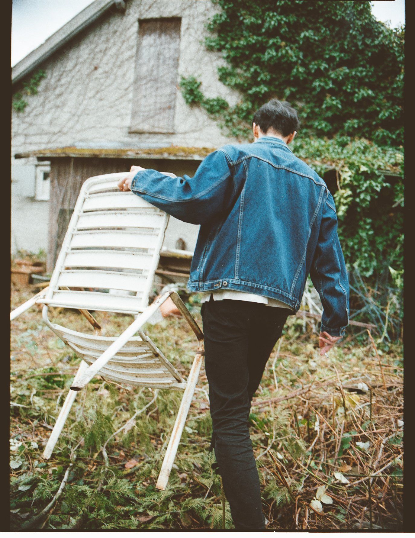Eine Person in einer Jeansjacke trägt einen weißen Stuhl in einem grasbewachsenen Hof mit einem Gebäude im Hintergrund.