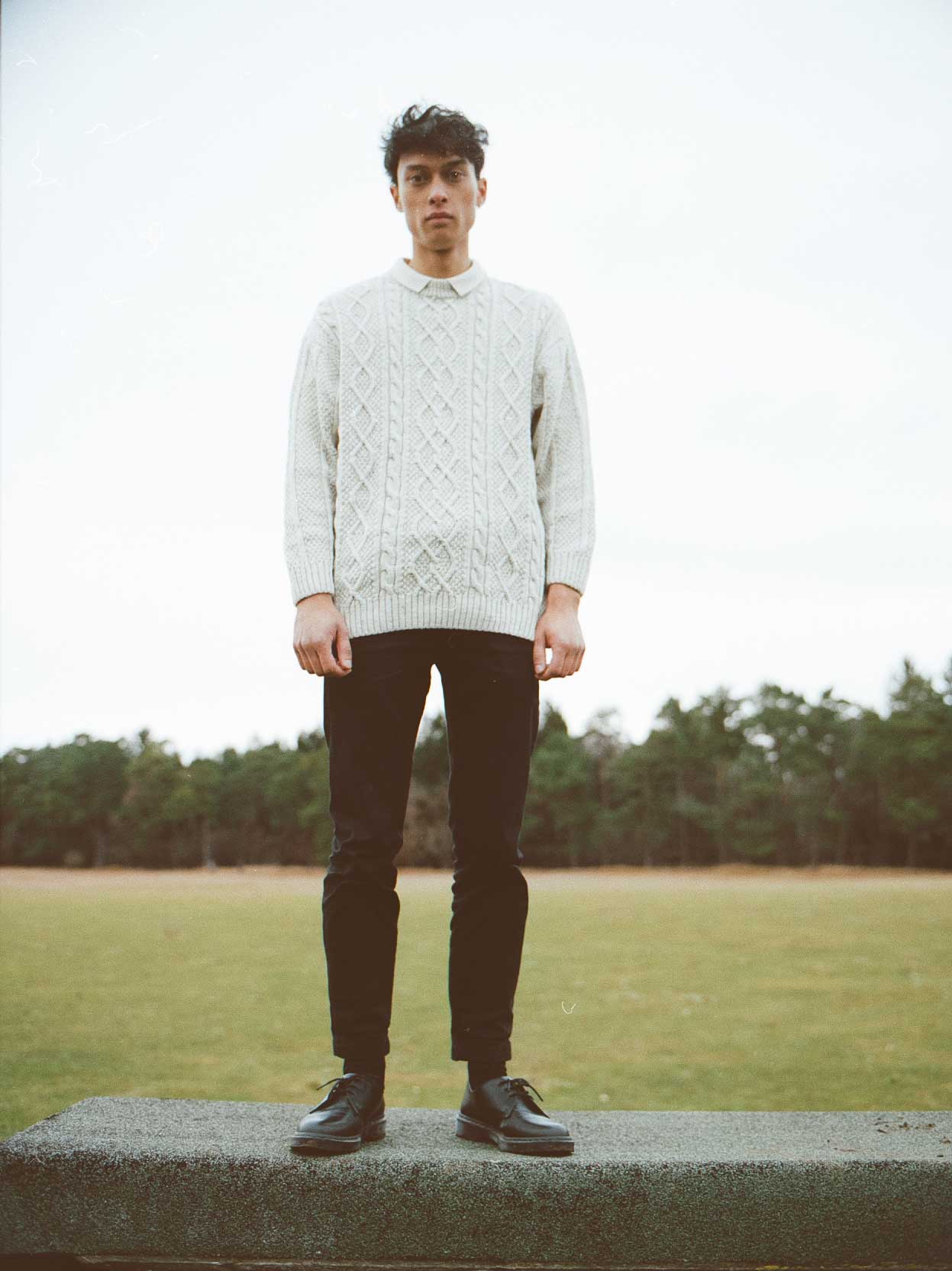 Mann in Zopfmusterpullover und schwarzer Hose steht im Freien auf einem Felsvorsprung, im Hintergrund grünes Feld und Bäume.