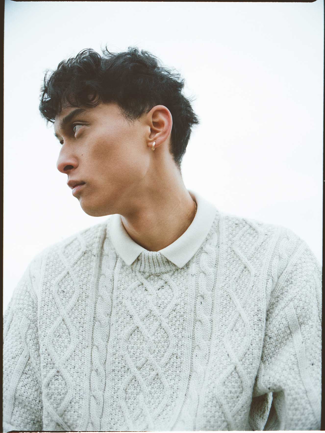 Mann mit lockigem Haar, hellgrauem Pullover, Blick nach links, im Freien.