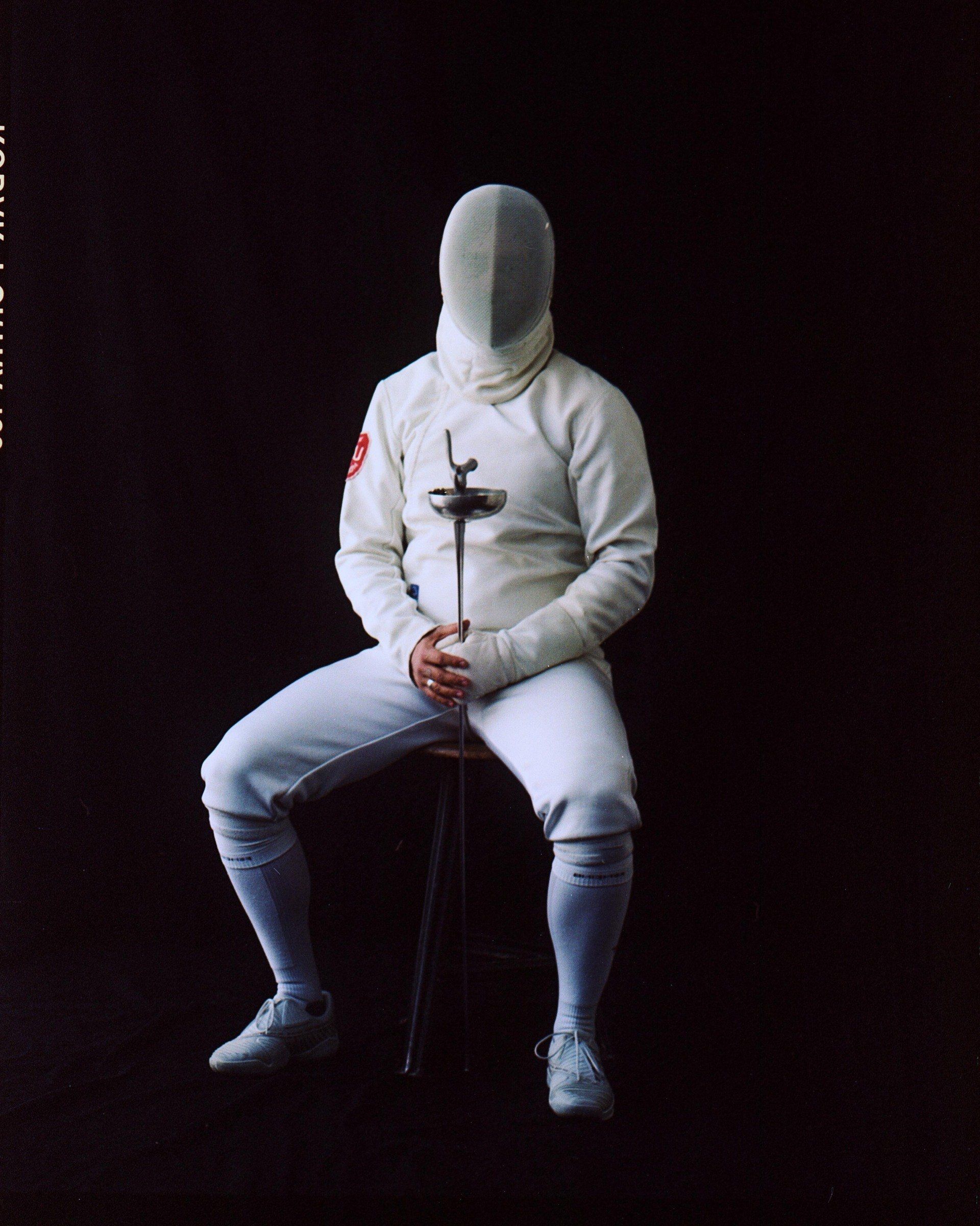 Fechter in weißer Uniform, sitzend mit Säbel, Gesicht verdeckt, auf schwarzem Hintergrund.