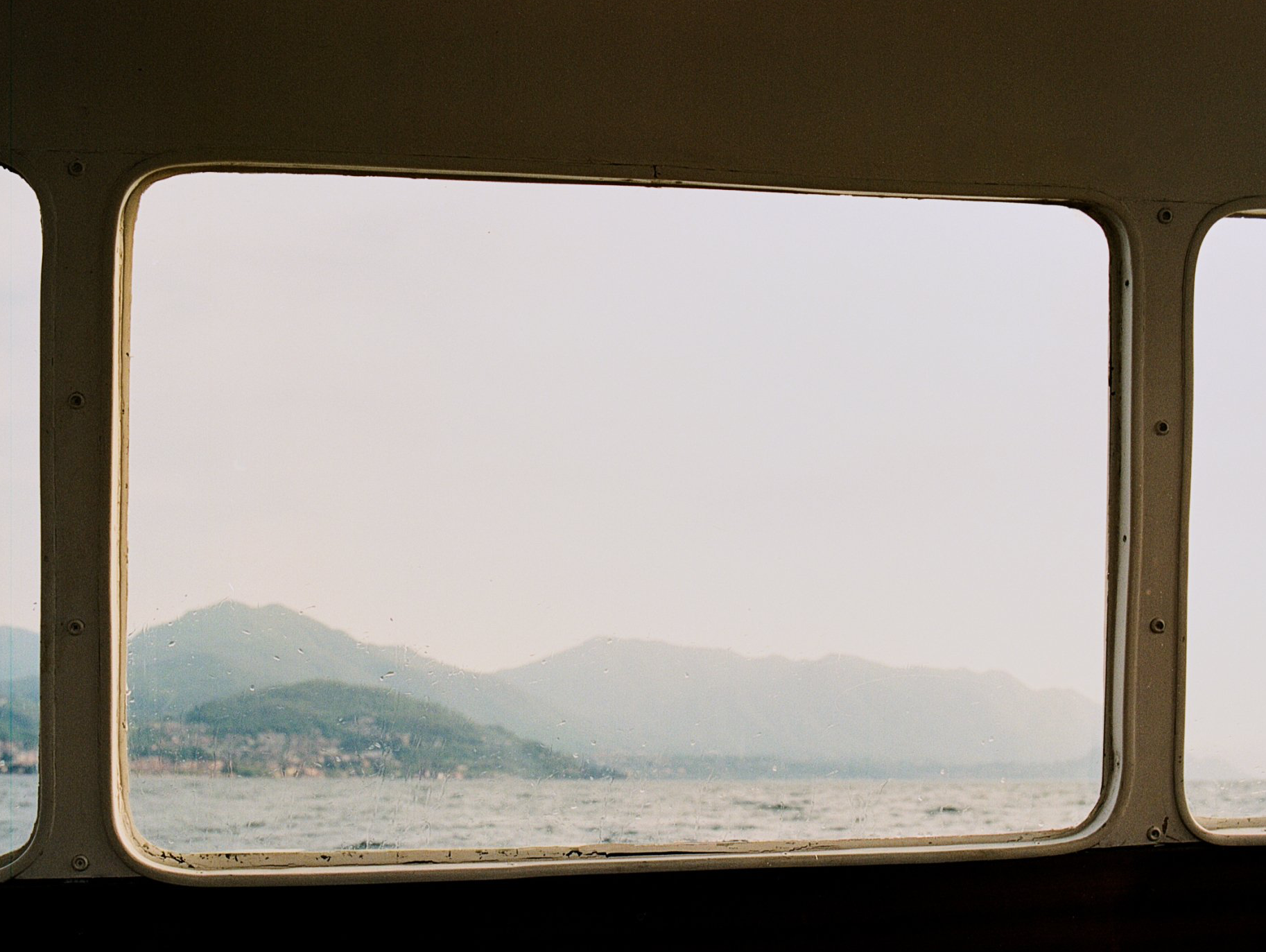 Blick aus einem Bootsfenster: Berge und Meer unter einem dunstigen Himmel.