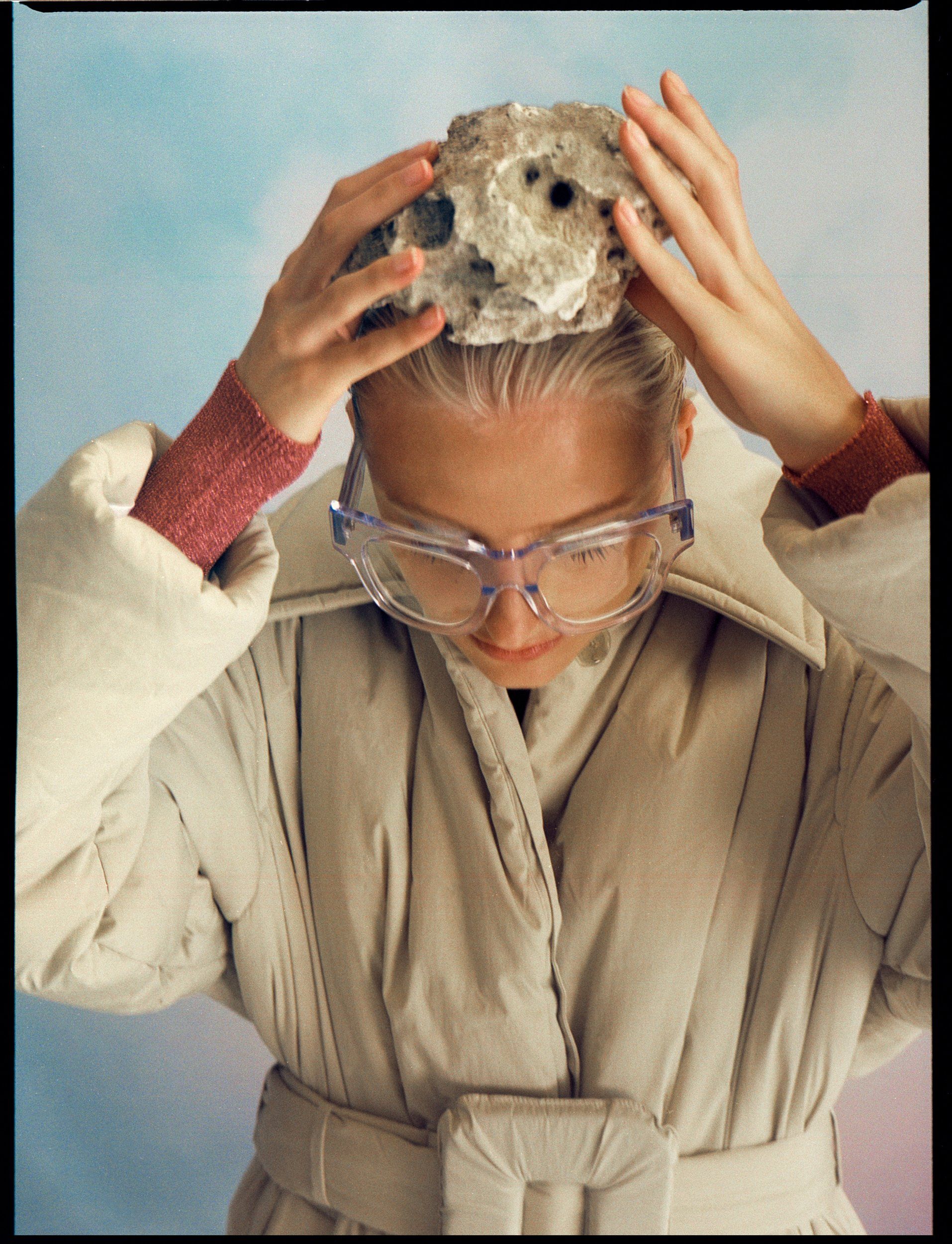 Frau mit Schutzbrille, hält einen Stein auf dem Kopf, heller Mantel, pastellfarbener Hintergrund.