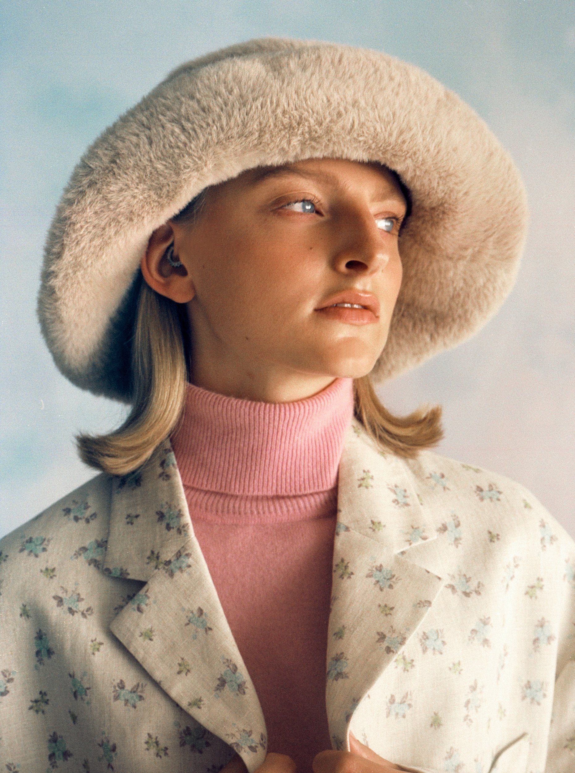 Frau mit Flauschmütze, rosa Rollkragenpullover und geblümtem Blazer, Blick zur Seite.