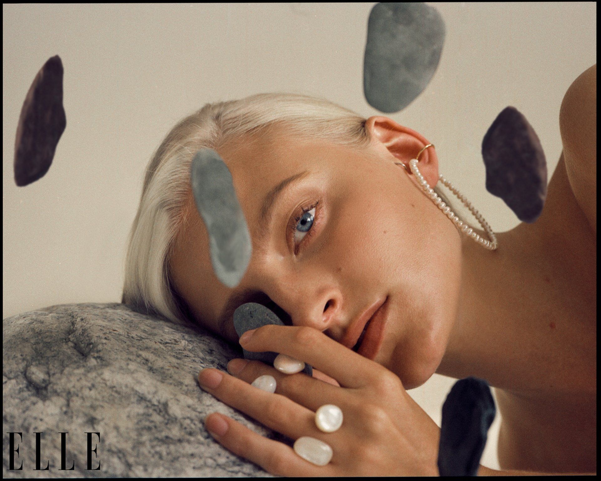 Blonde Frau mit blauen Augen, Perlen und Steinen. Sie liegt auf einem Felsen, um ihr Gesicht schwimmen Steine.