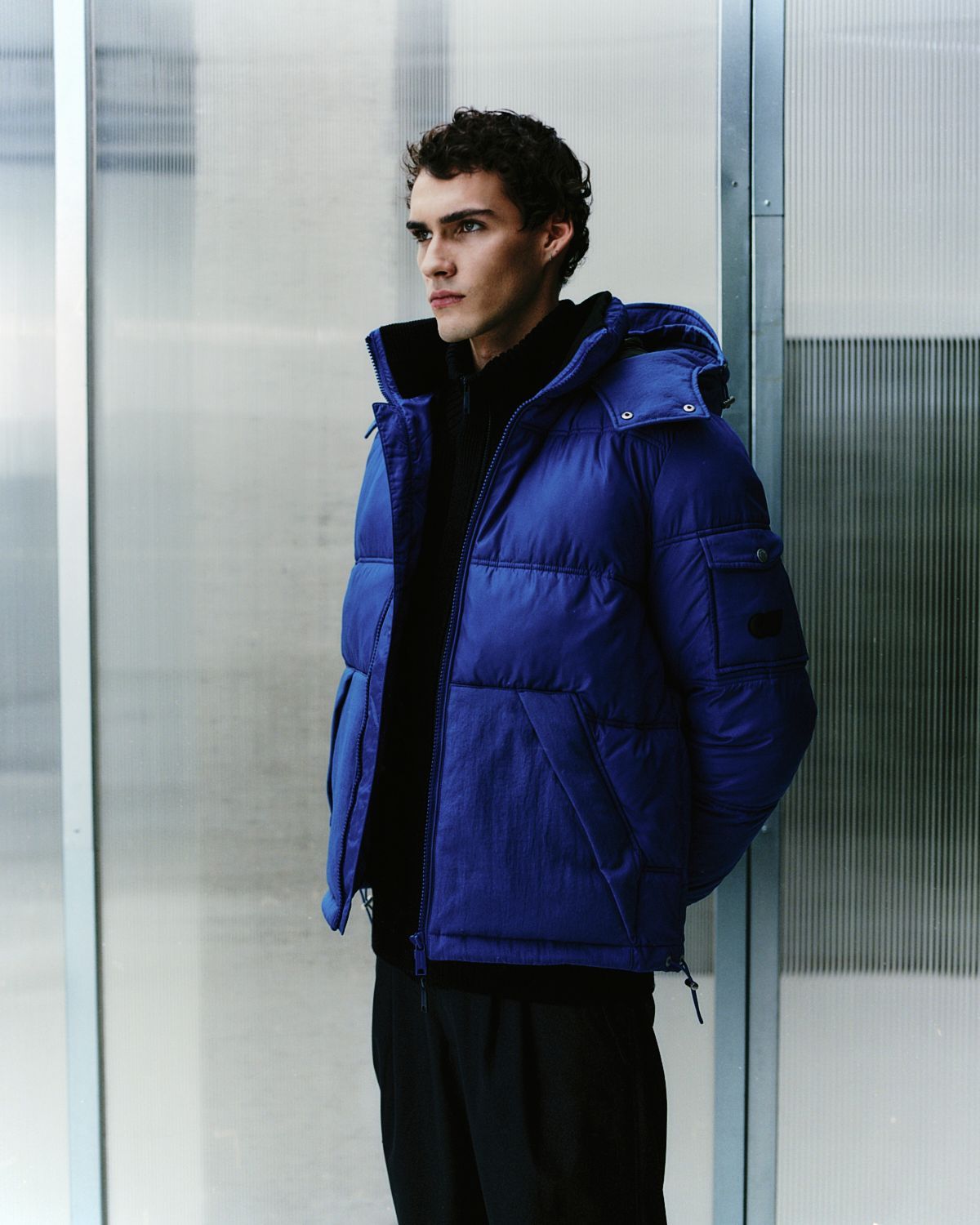 Mann in blauer Pufferjacke und schwarzer Kleidung steht an einer modernen Wand und blickt zur Seite.