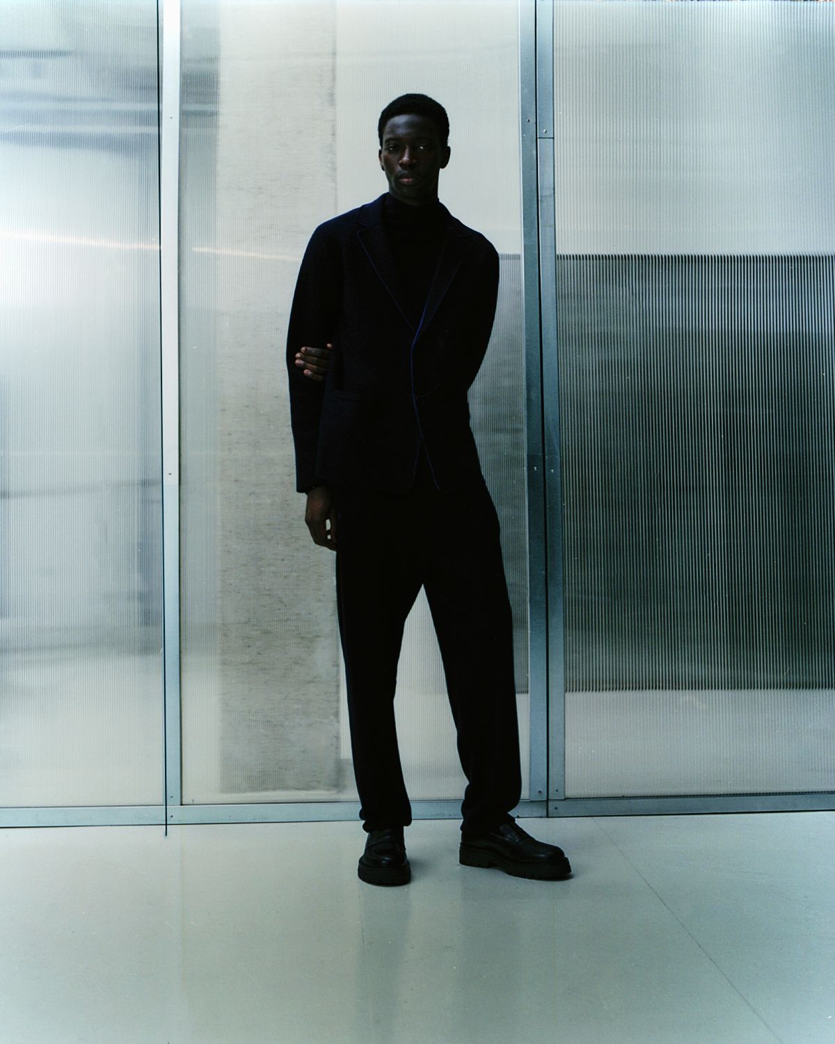 Mann im schwarzen Overall posiert vor gemusterter Glaswand.