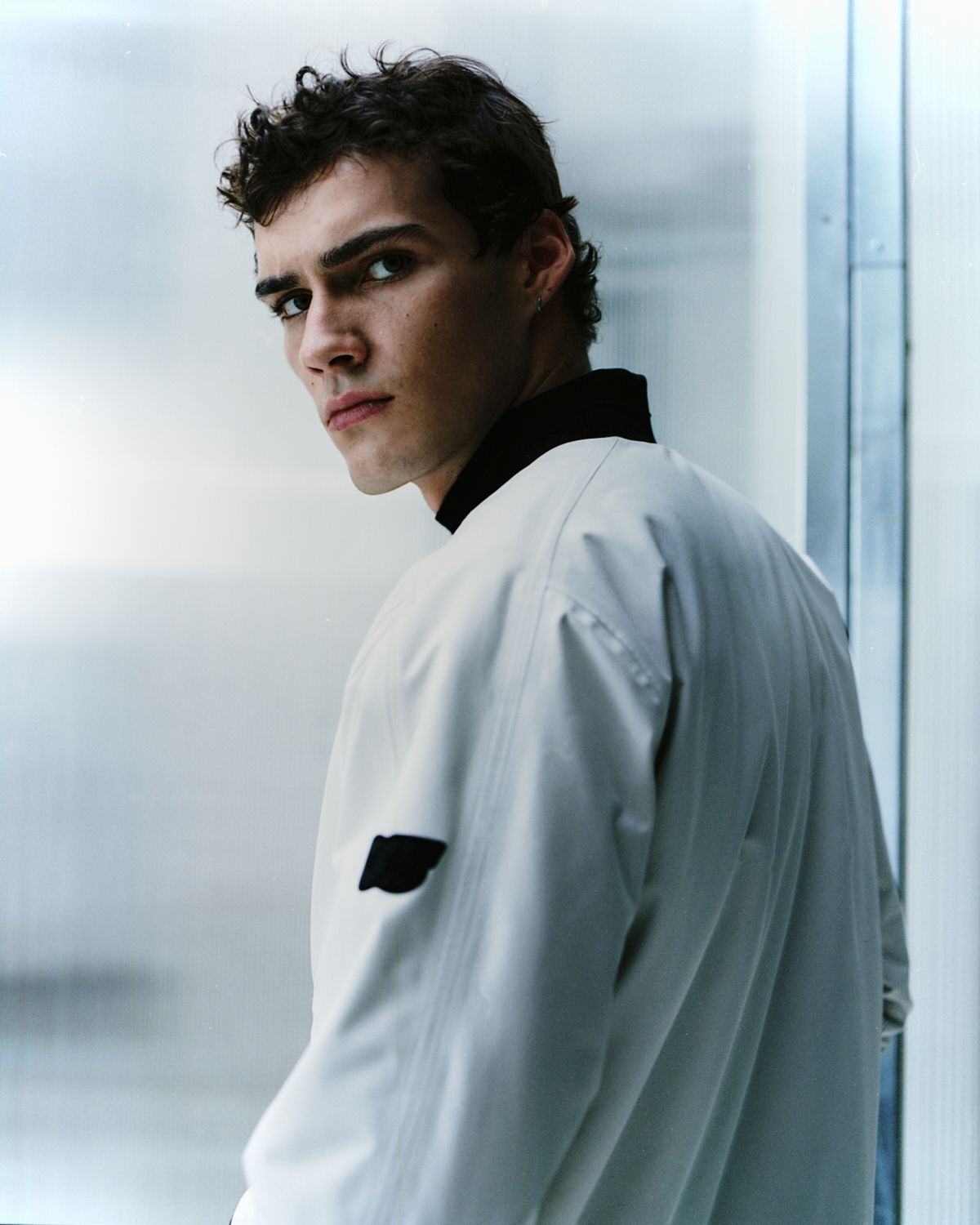 Mann mit dunklem lockigem Haar schaut über seine Schulter, trägt weiße Jacke und schwarzen Kragen.