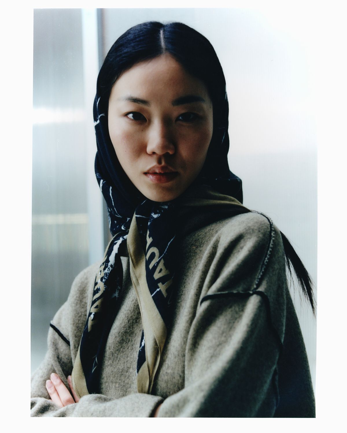 Frau mit dunklem Haar und Schal, trägt einen hellen Pullover, die Arme verschränkt und blickt den Betrachter an.